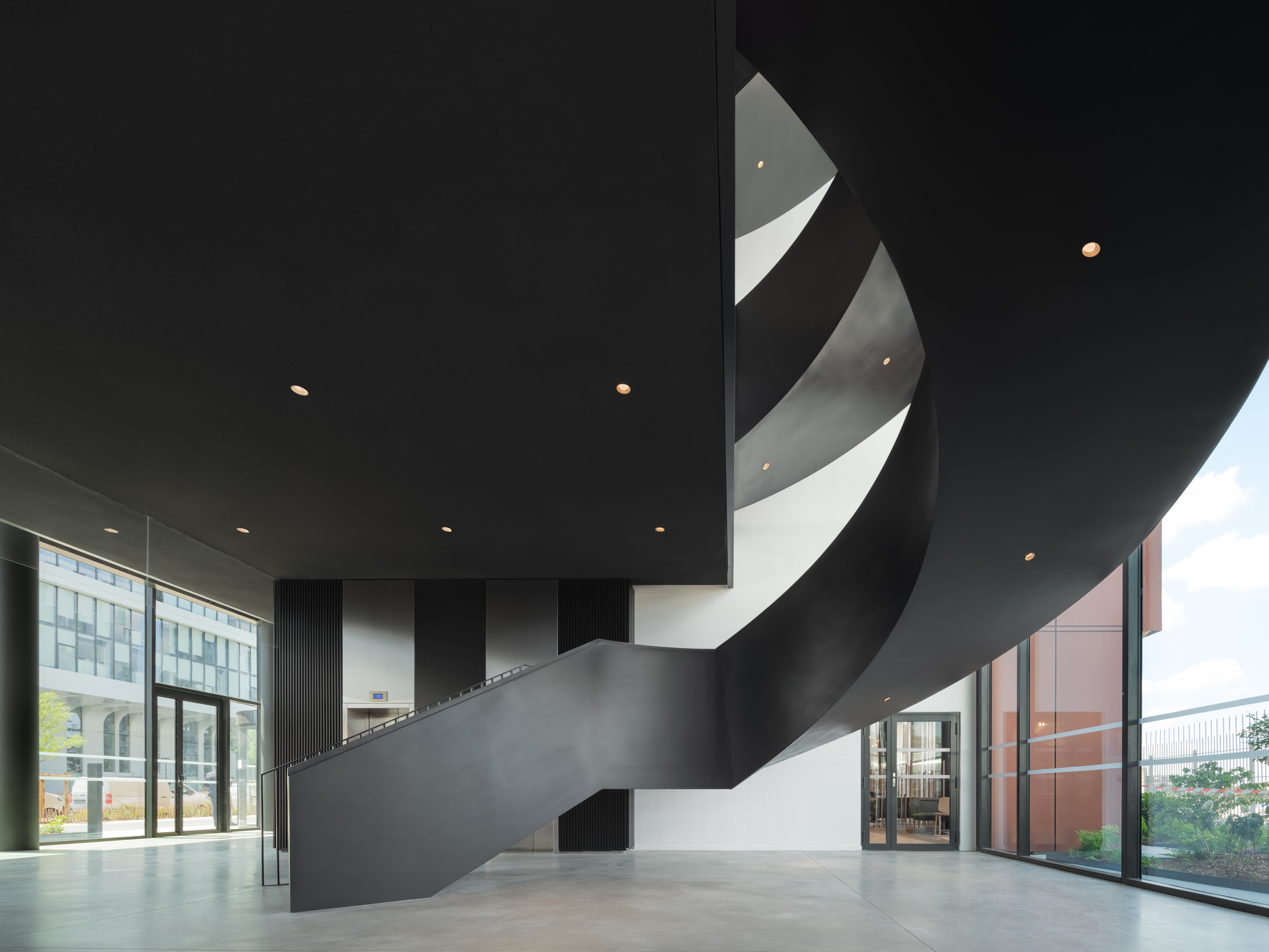 Interior lobby with black ribbon staircase spiraling beneath recessed ceiling downlights and glazed walls