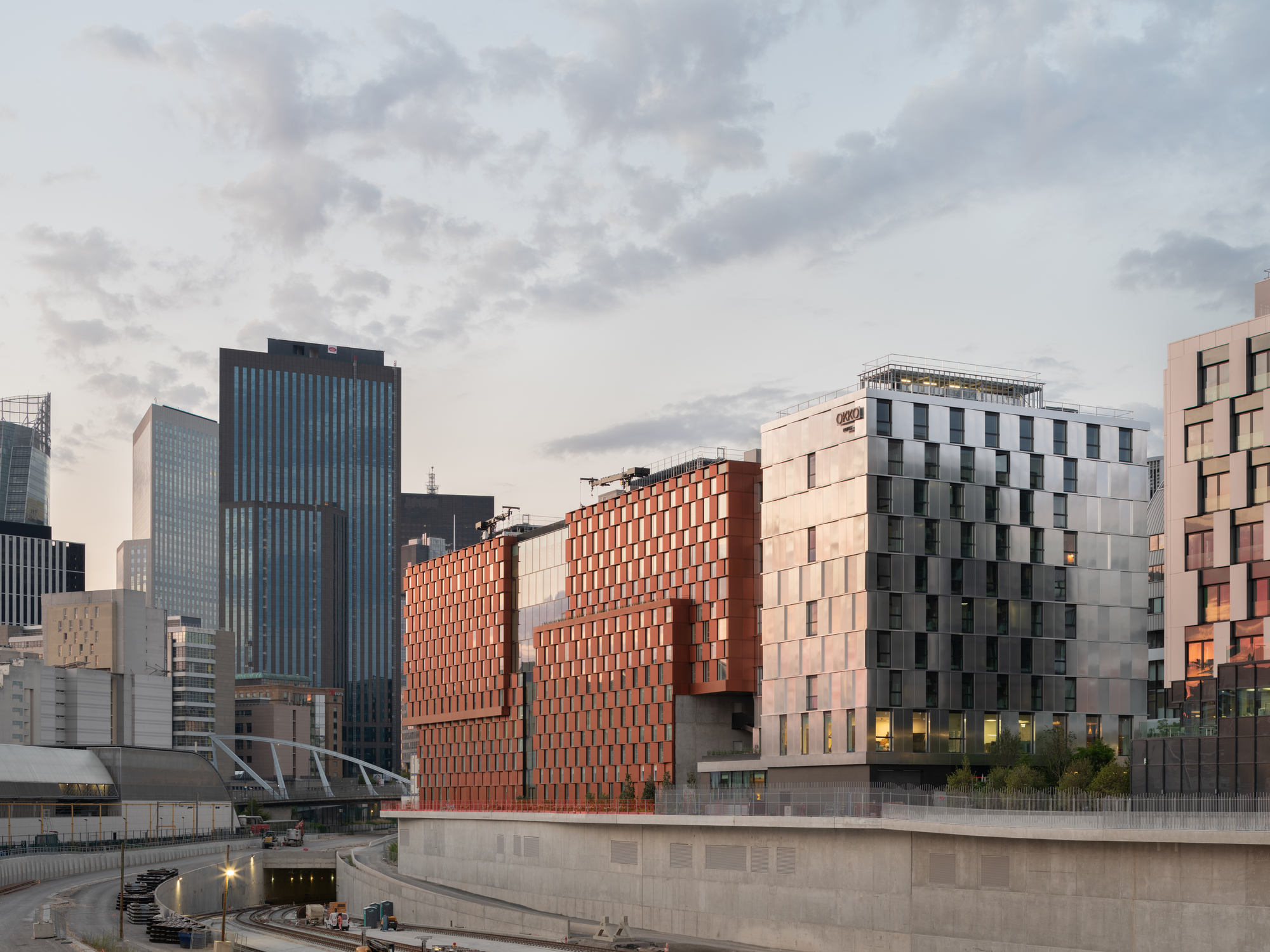 Urban context showing the red paneled volumes among neighboring towers at sunset beside a water feature