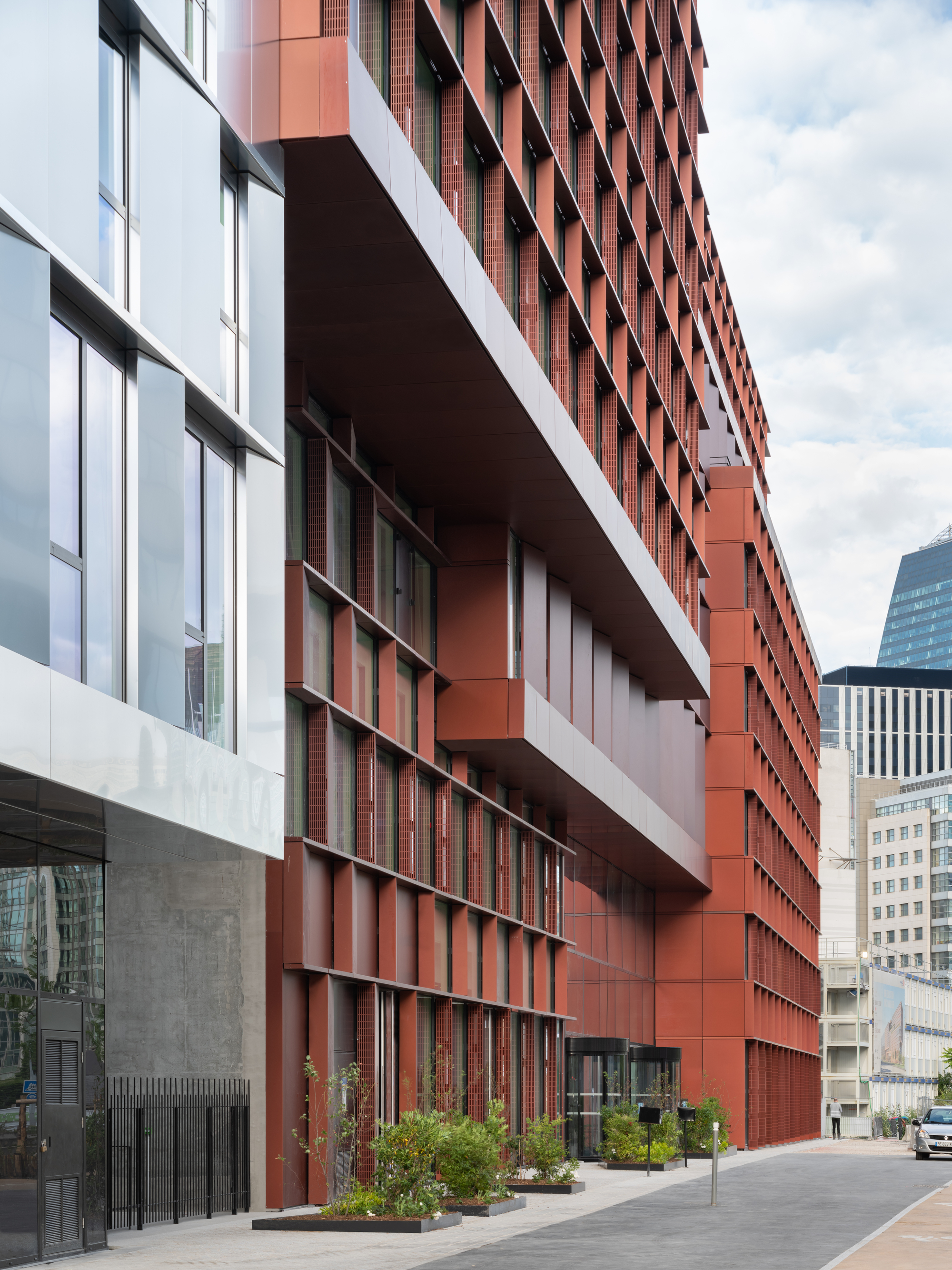 Street-level view of stacked terracotta volumes above a concrete base with planted beds and pedestrians