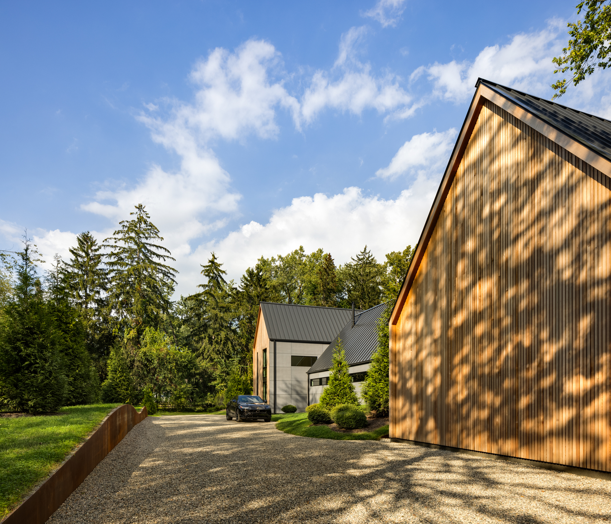 Gravel motor court flanked by timber-clad volumes with a parked vehicle and scattered tree shadows