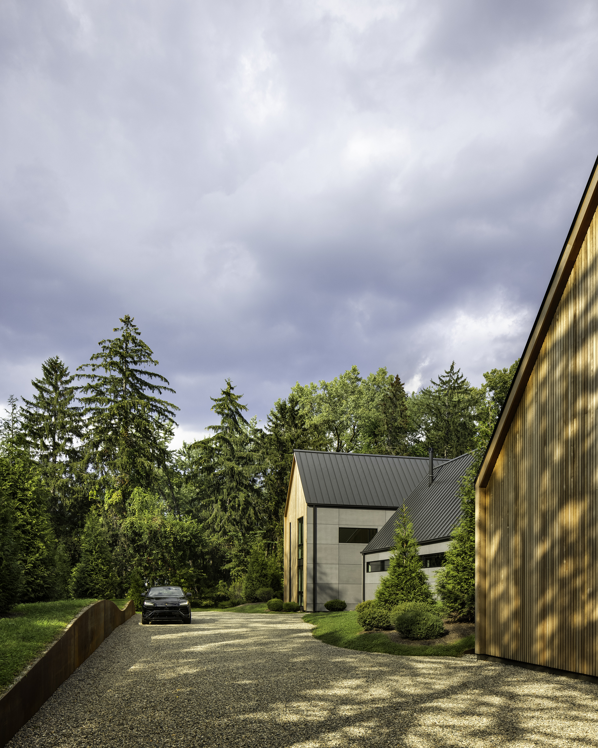 Gravel driveway approach showing white and timber volumes with standing-seam metal roofs under overcast skies