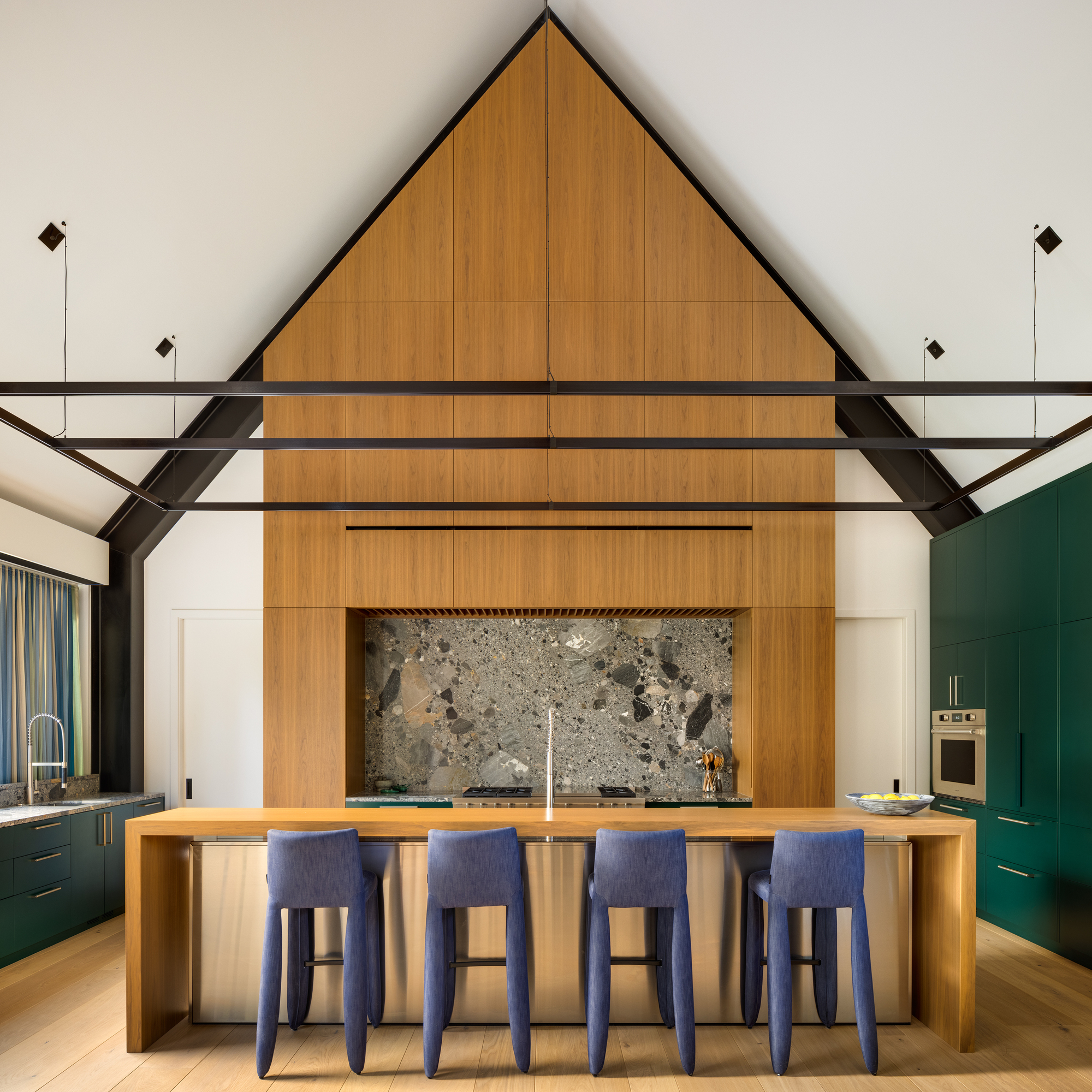 Kitchen island with four blue upholstered stools beneath timber-clad gable wall with terrazzo backsplash