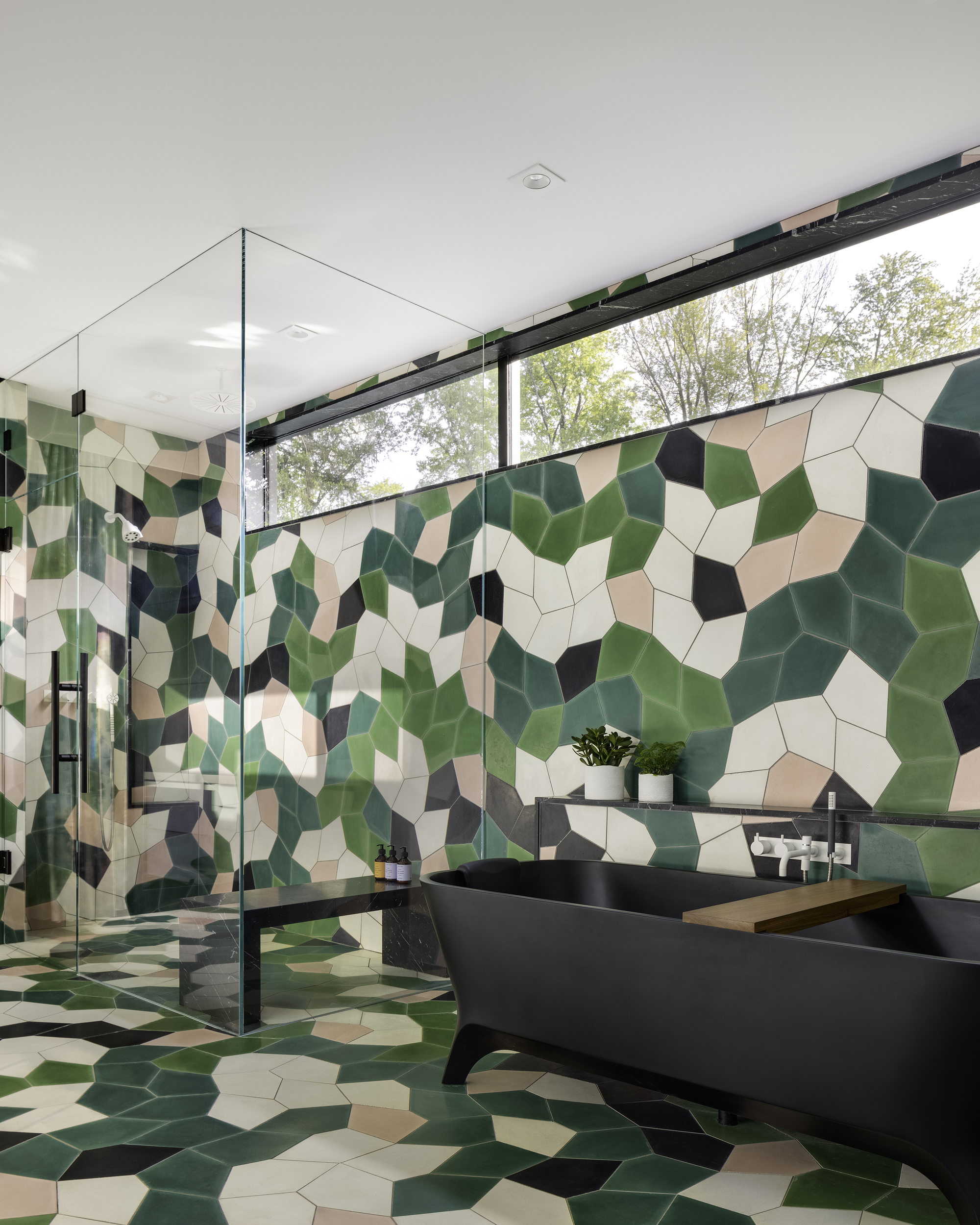 Bathroom with camouflage-patterned mosaic tile walls and floor, glass shower enclosure, and clerestory window overlooking trees
