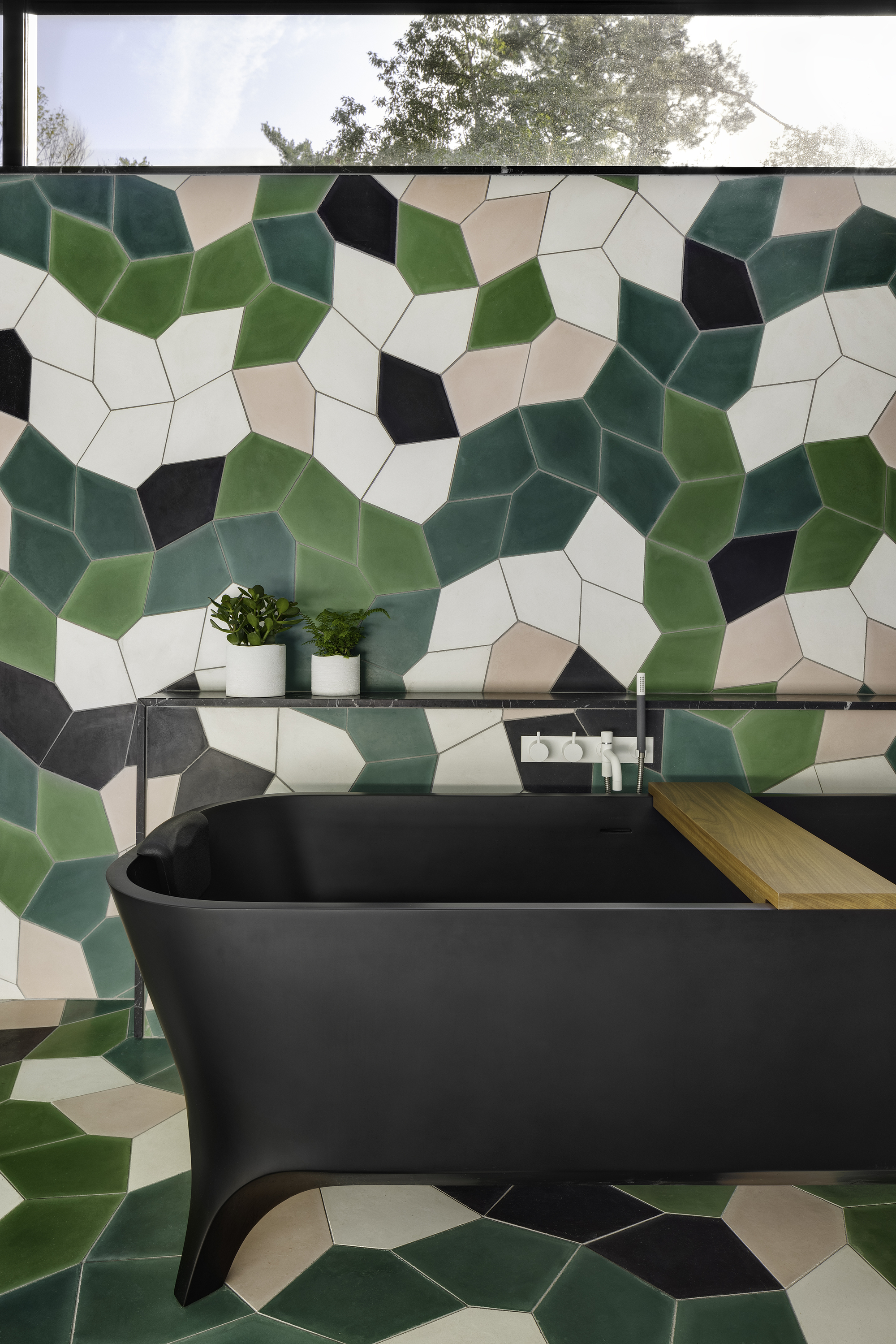 Black freestanding bathtub with timber shelf against geometric tile wall in green, black, and cream tones