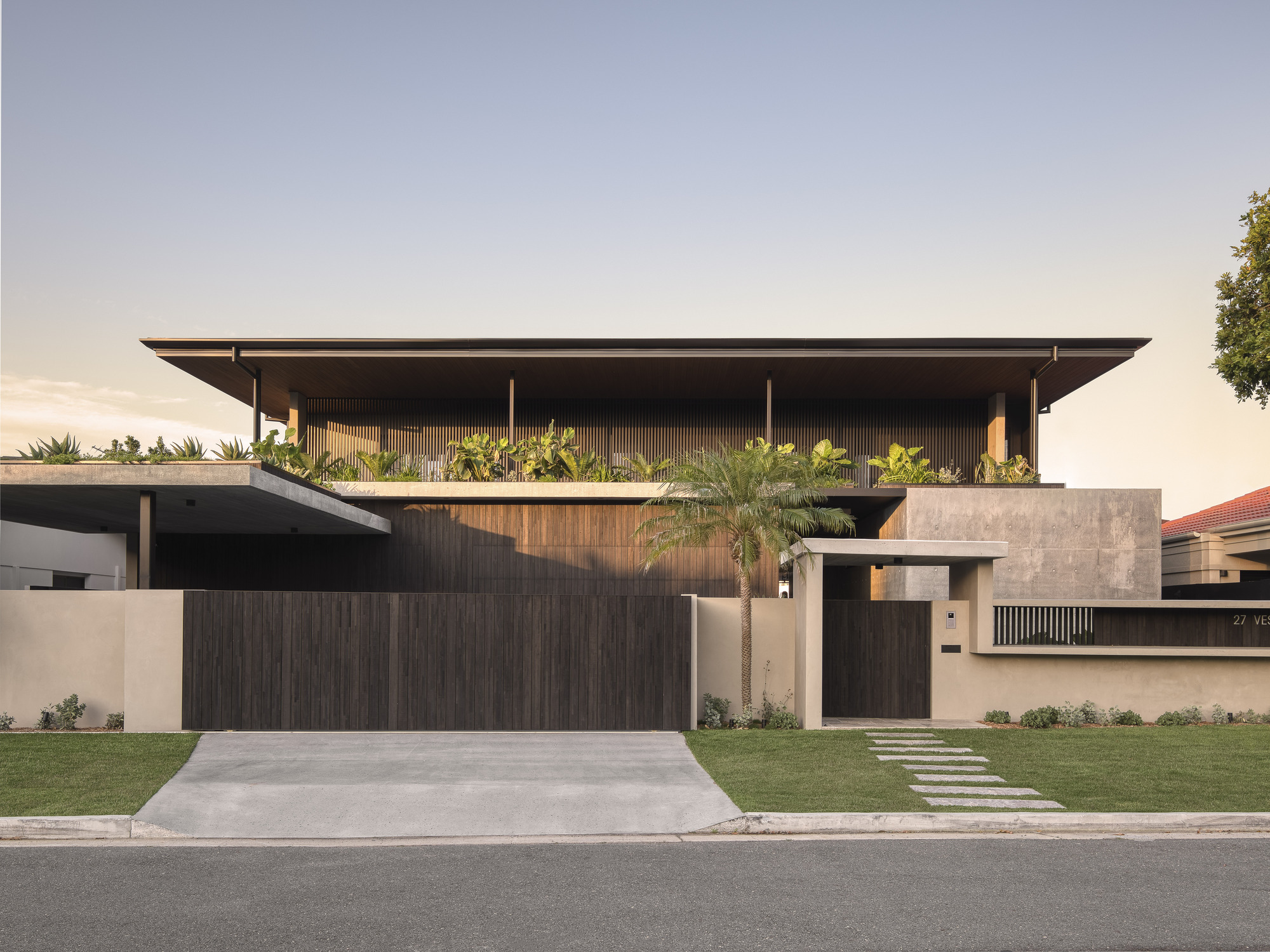 Street view of the two-level facade with timber cladding, concrete walls and planted upper terrace at dusk