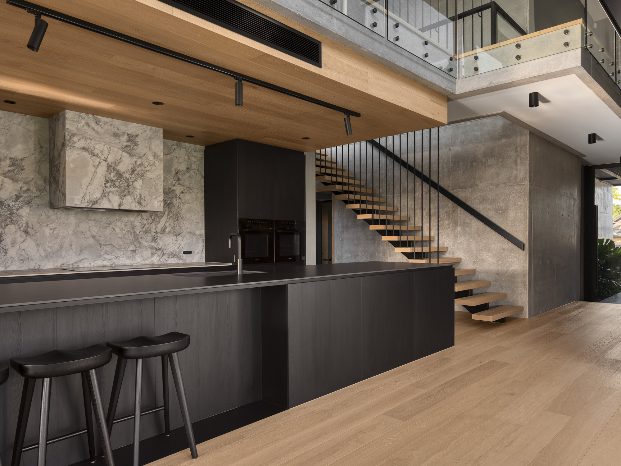 Double-height interior showing floating timber staircase with vertical steel balusters against exposed concrete walls