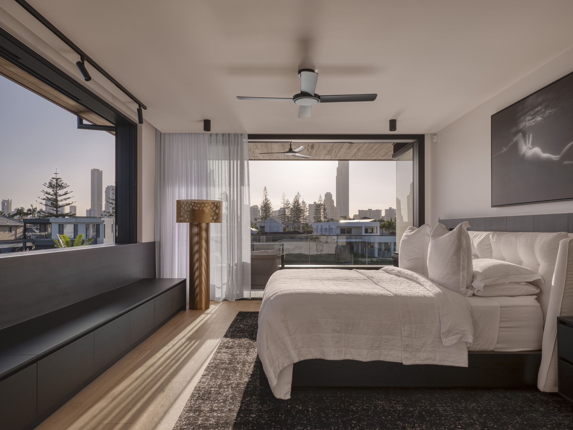 Bedroom with corner glazing and balcony doors framing views of residential rooftops at twilight