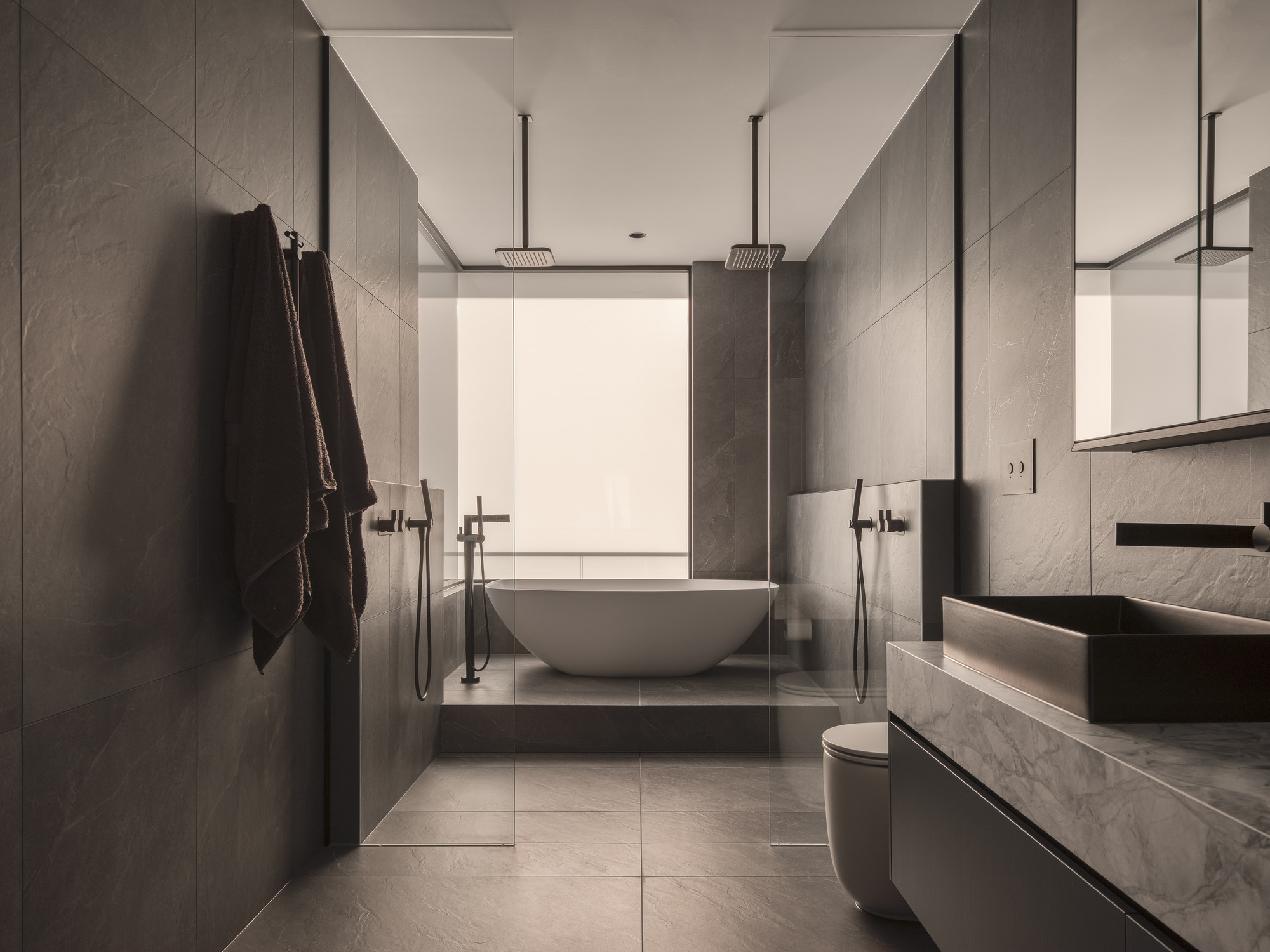 Bathroom with grey stone panels, freestanding tub, glass shower enclosure and dual rainfall showerheads
