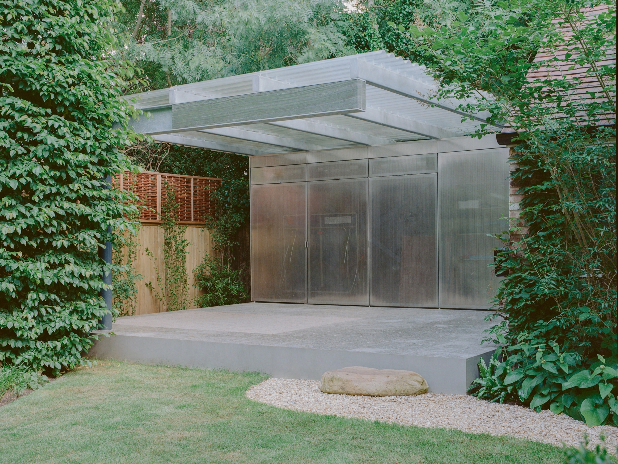 Concrete porch with gravel path leading to the translucent metal entry pavilion under a cantilevered roof