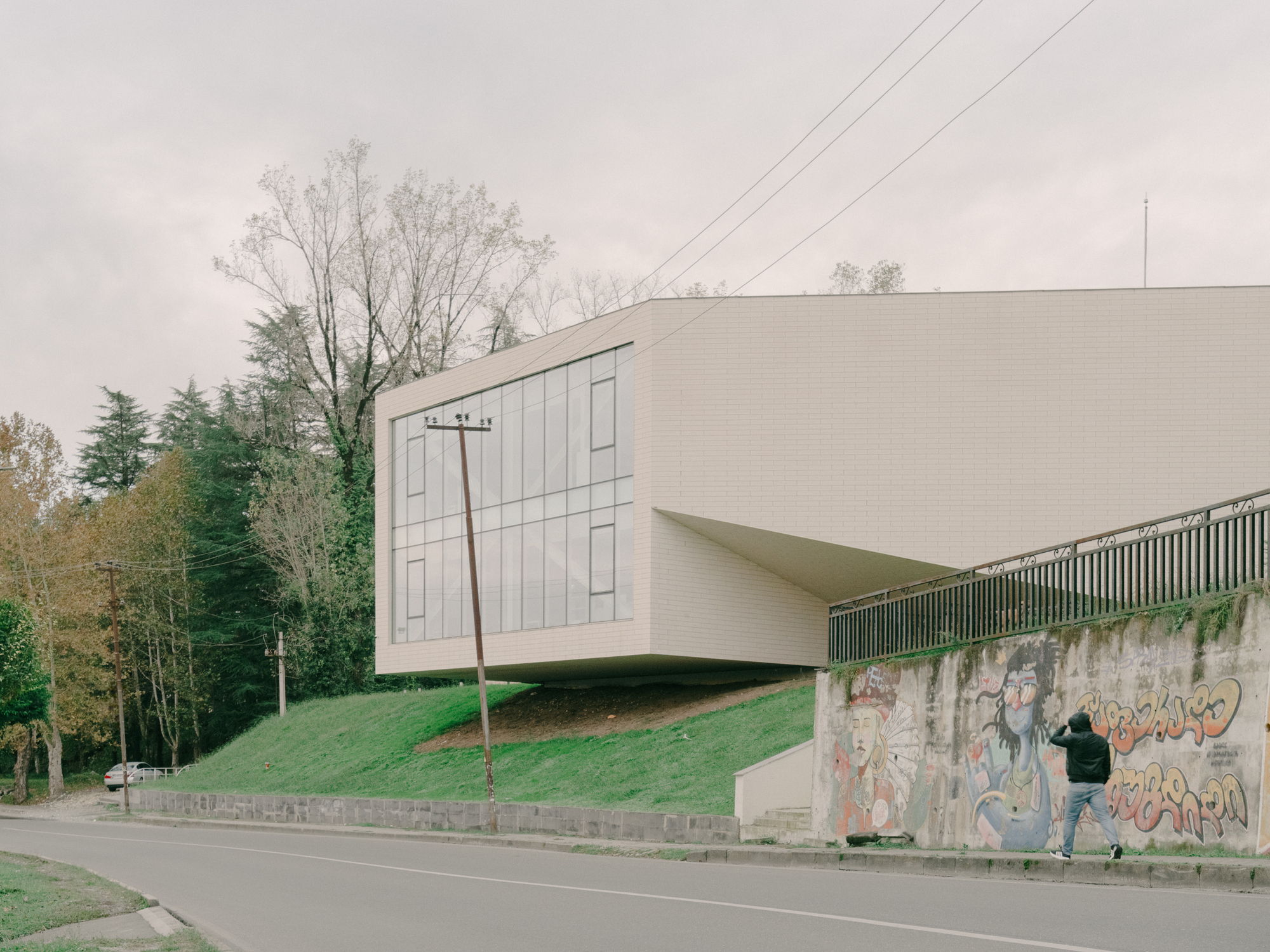 Street view of the elevated white volume above a graffitied retaining wall with a person walking below