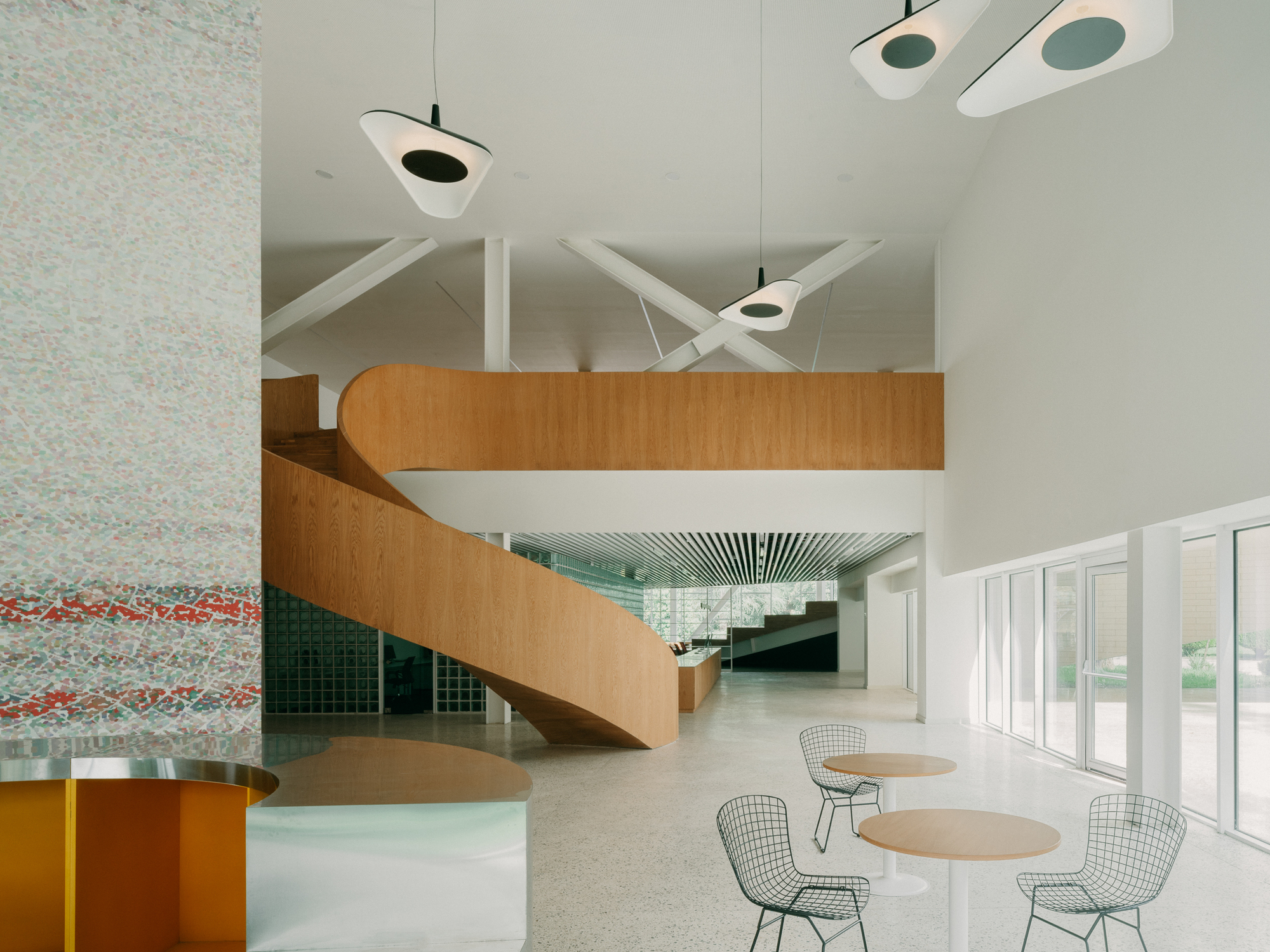 Double-height lobby with curving timber staircase, pendant lights, and wire mesh chairs at round tables