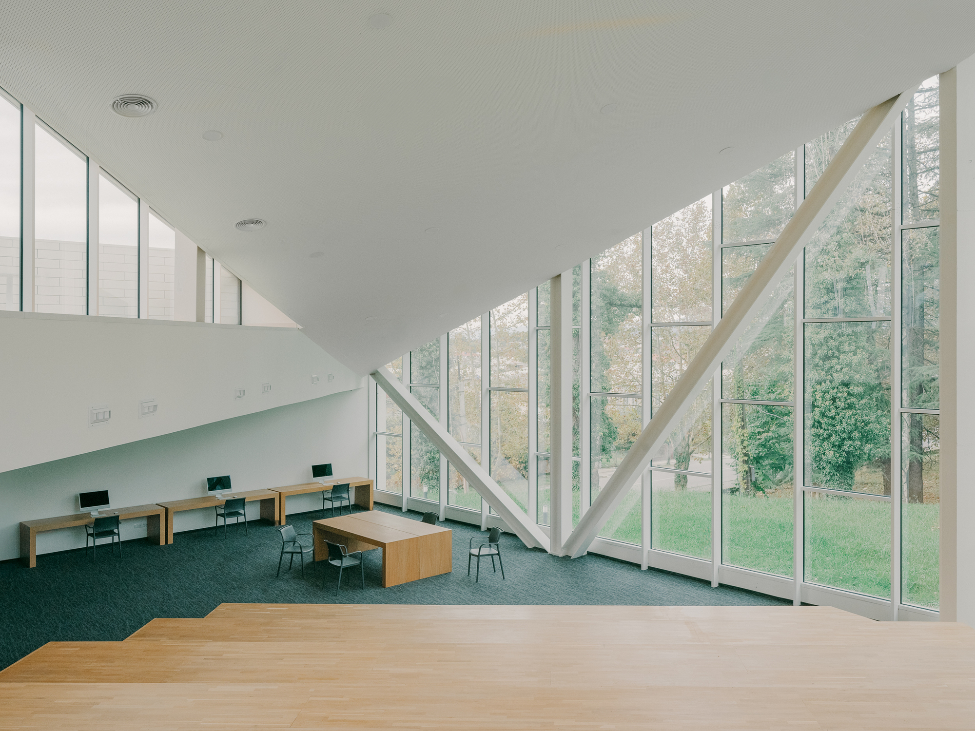 Double-height reading room with angled white structural beams framing floor-to-ceiling windows overlooking a park