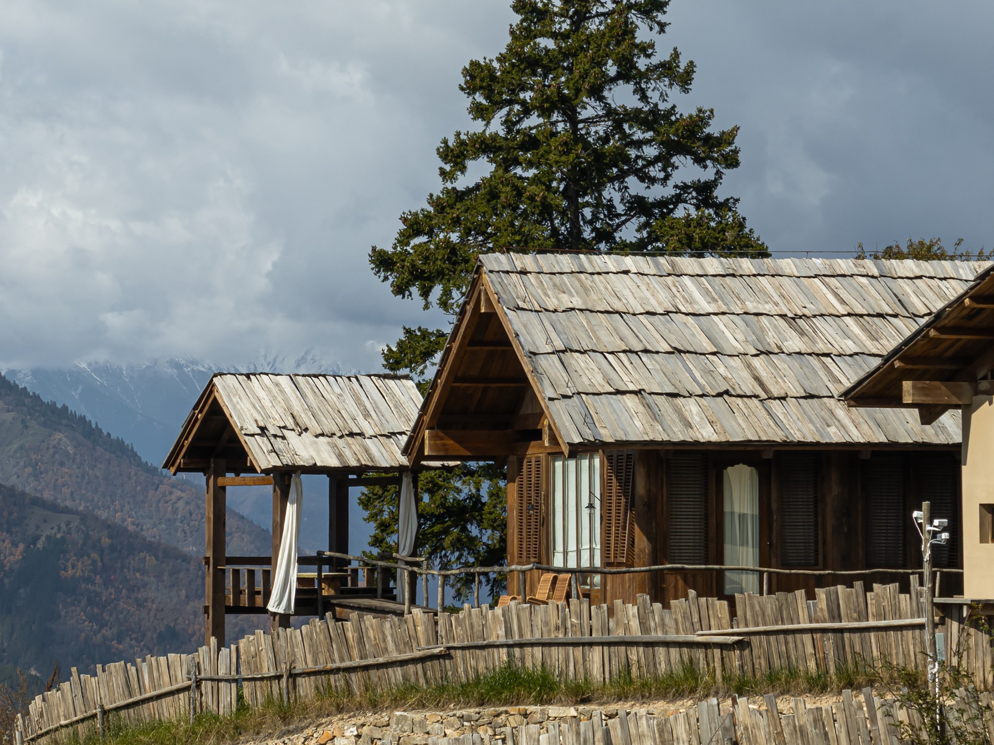 Pavilion with open timber frame and shingled roof beside dwelling units with slatted wood shutters under clouded sky