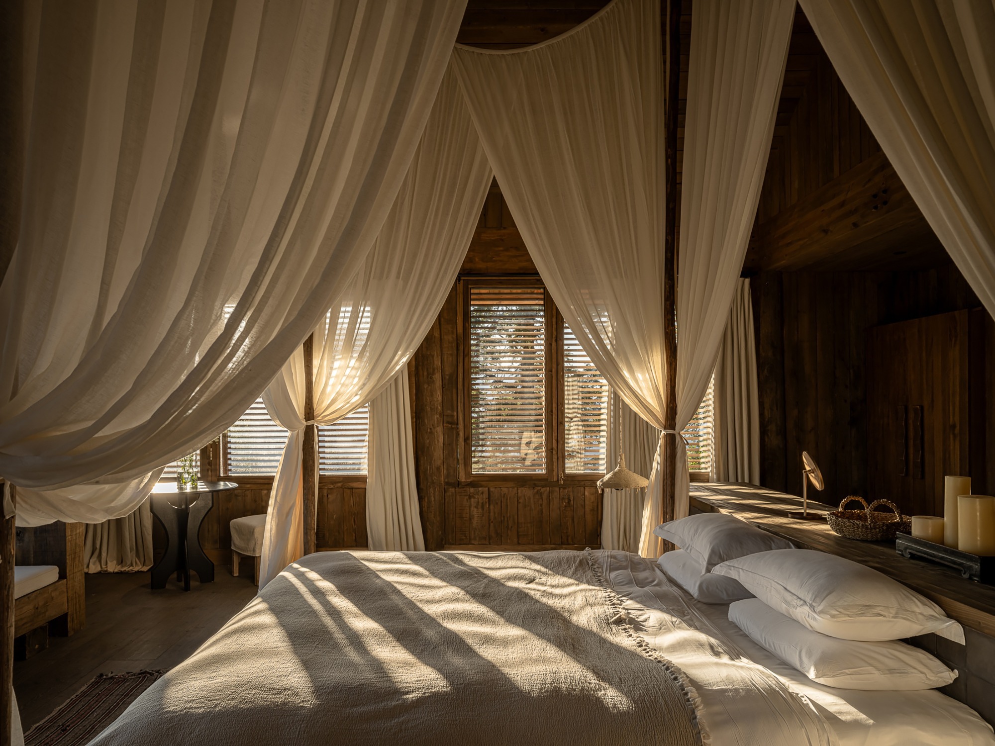 Bedroom with canopy bed draped in sheer curtains and timber louvers filtering sunlight onto the bedding