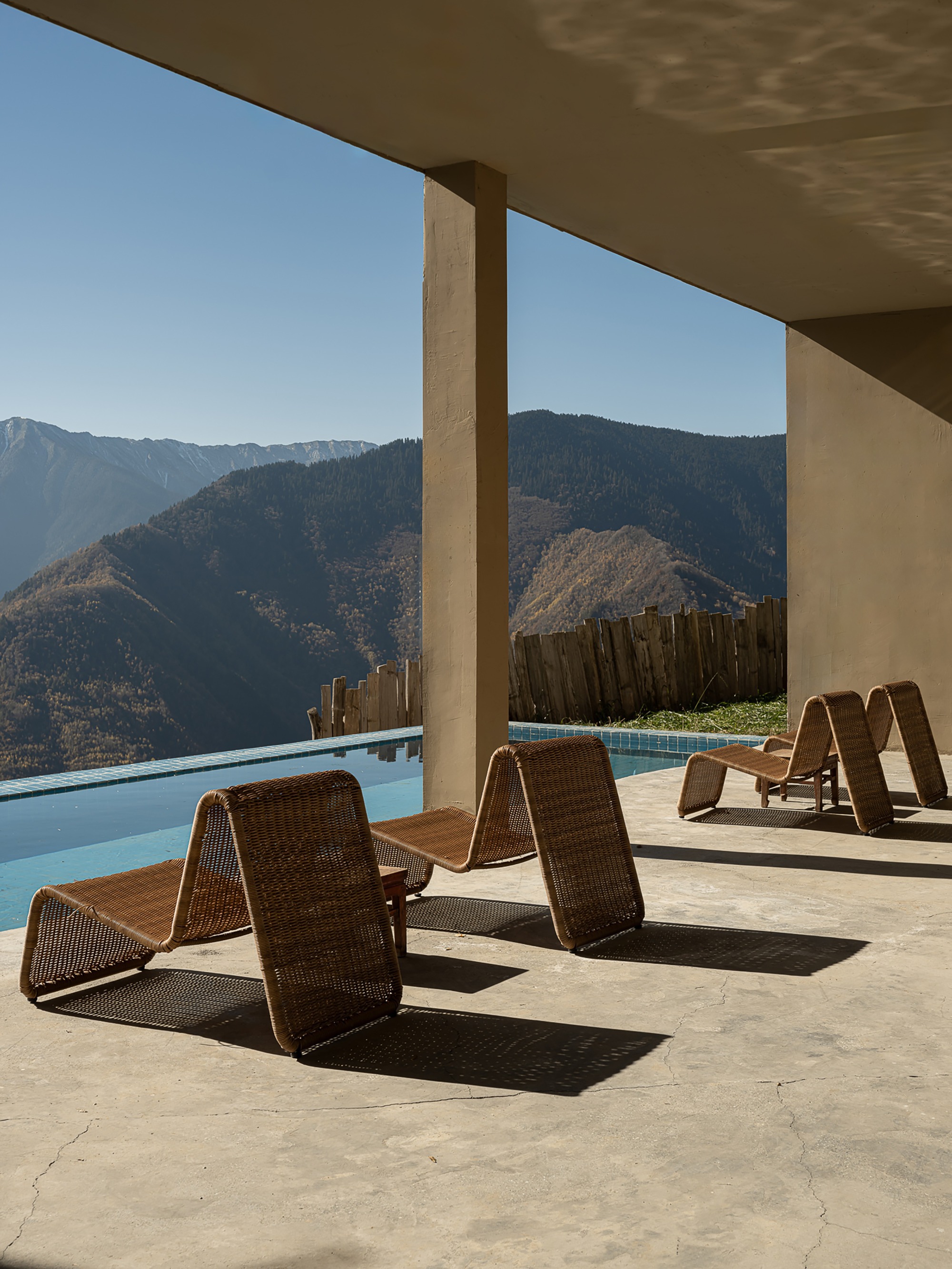 Covered terrace with woven lounge chairs and pool overlooking forested mountain peaks under clear sky