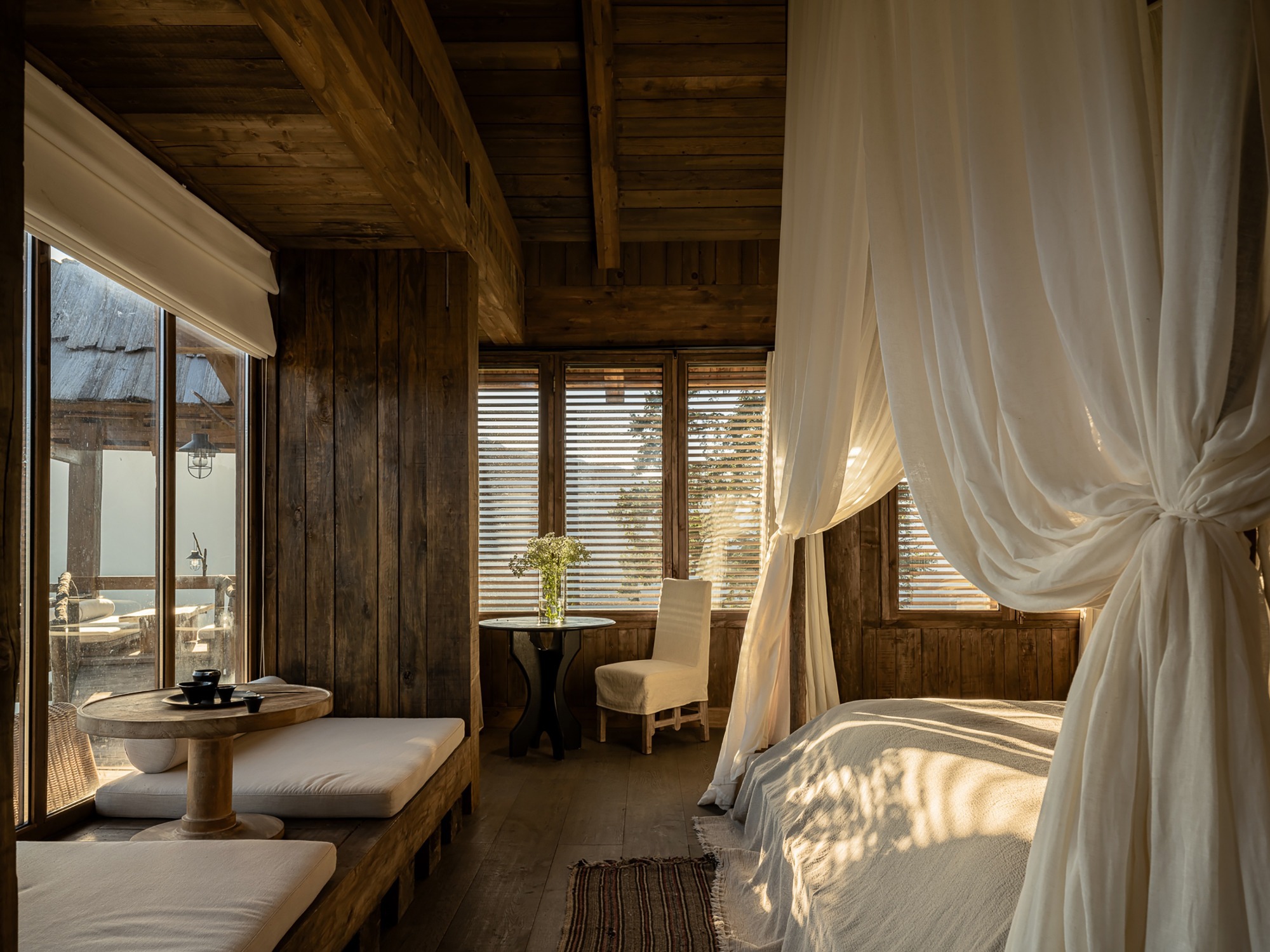 Spa room with timber walls, sheer canopy, treatment bed, and slatted windows filtering dappled light
