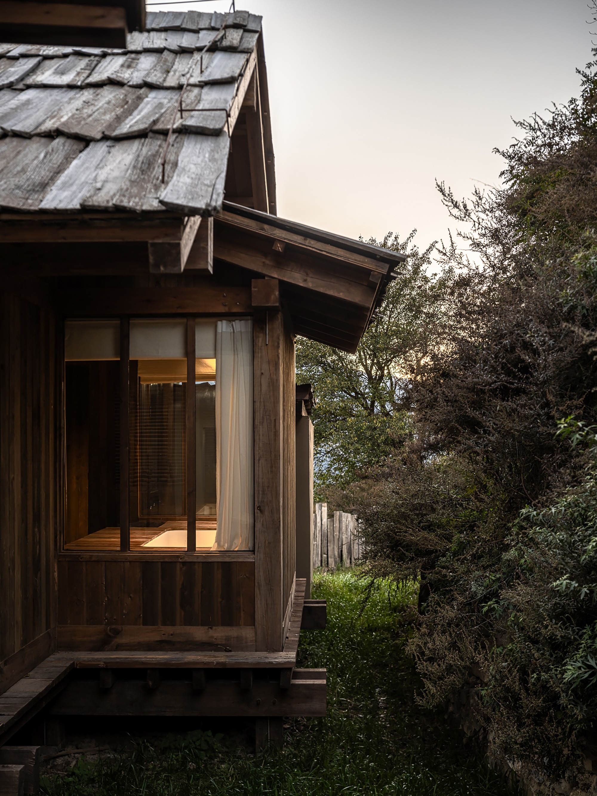 Timber-clad facade with weathered shingle roof and glazed corner bay at dusk