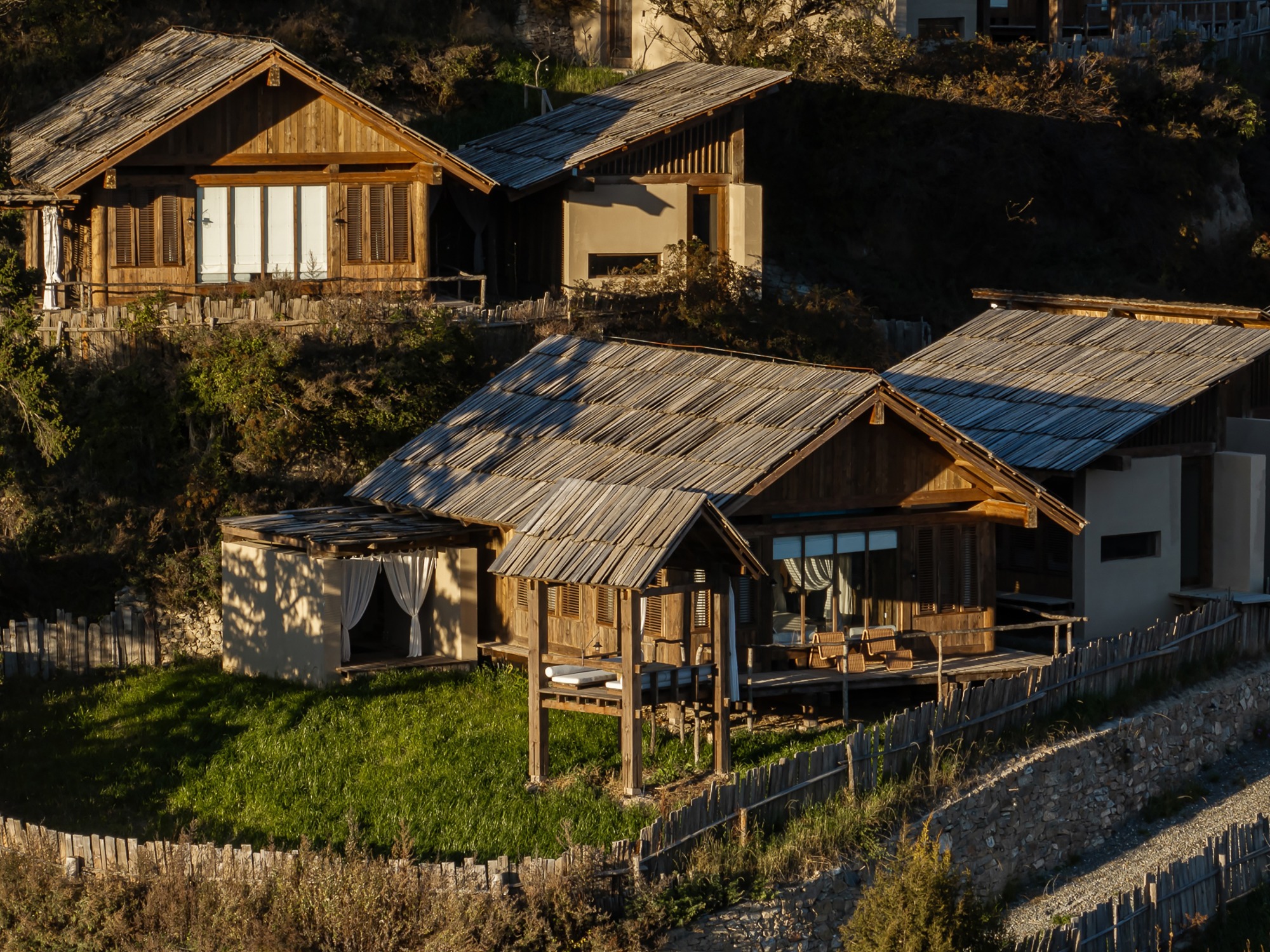 Cluster of timber and stone-clad volumes with corrugated metal roofs on a sloping green hillside