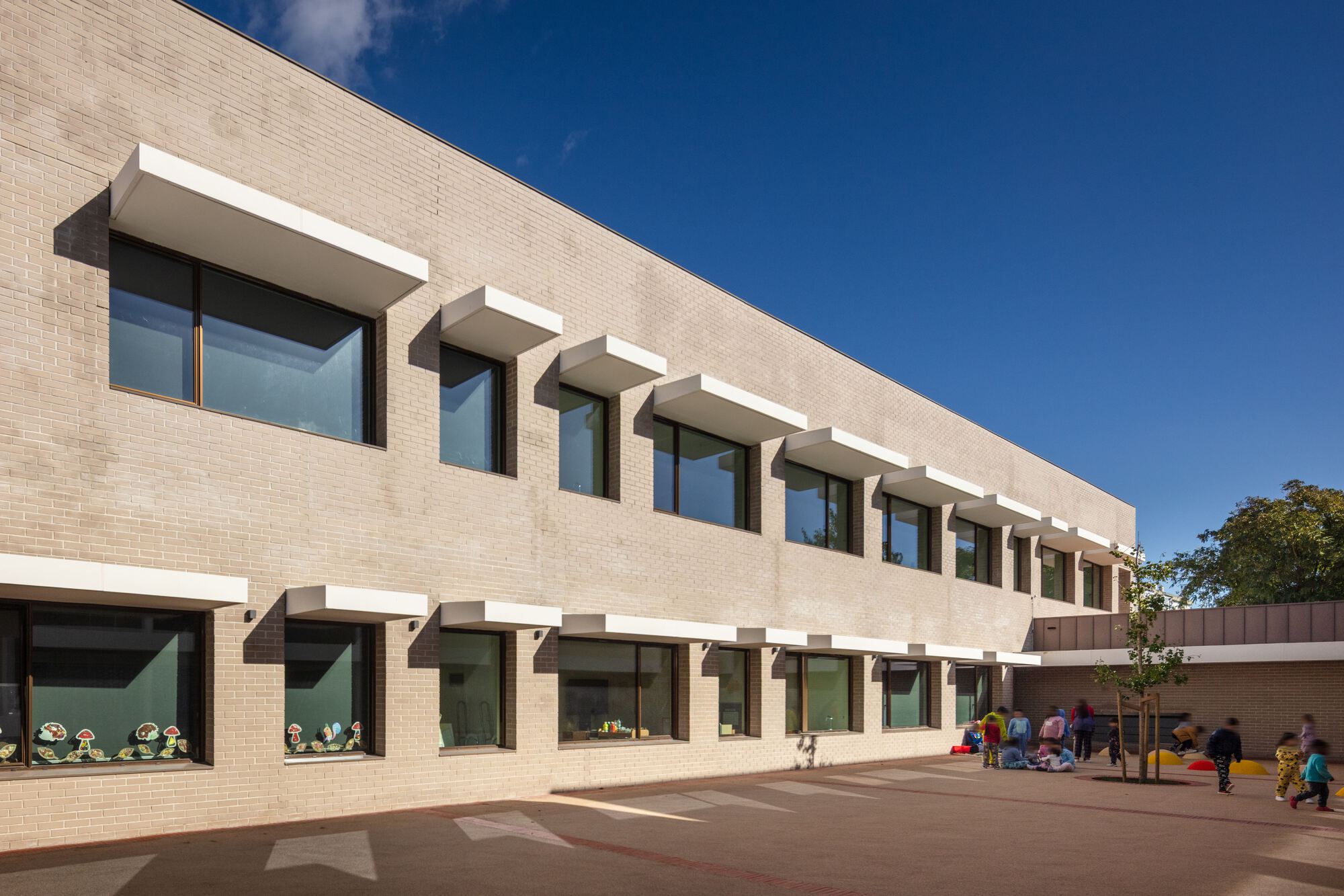 Courtyard facade with repeating canopy shades over glazed openings and children playing below