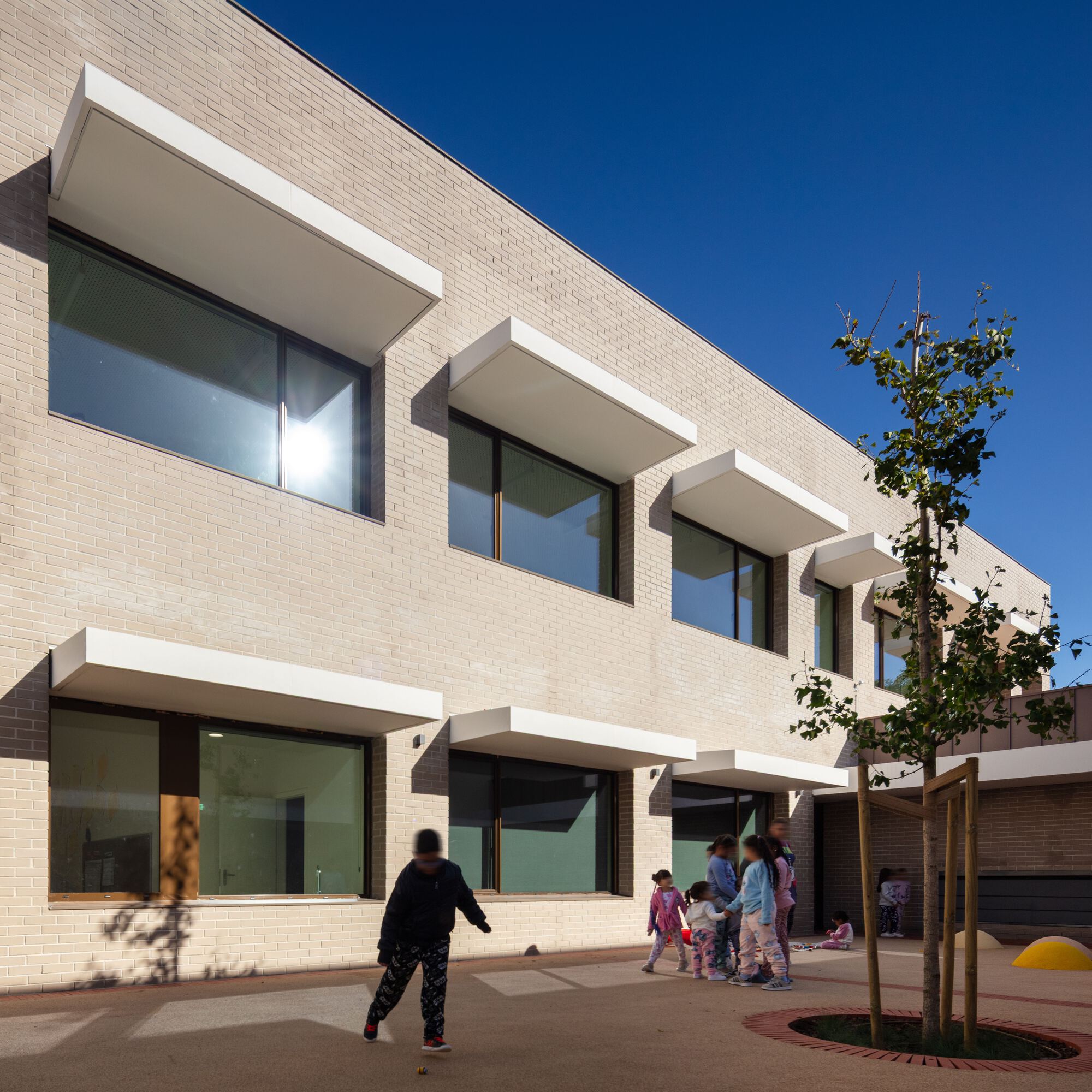 Two-story brick facade with projecting sun shades as children run across the paved courtyard