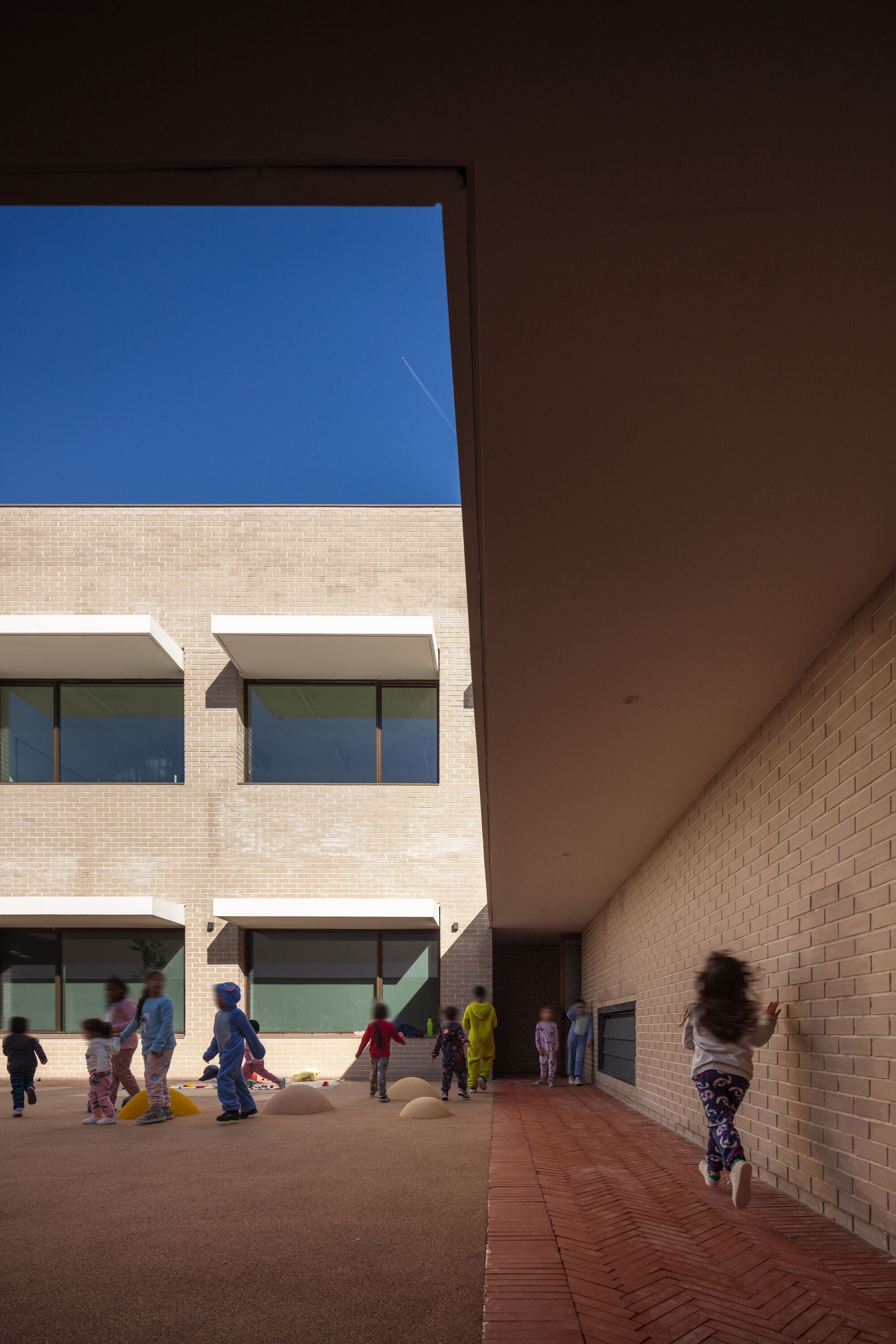 Covered walkway with deep overhang framing the brick facade and children moving through dappled shadow