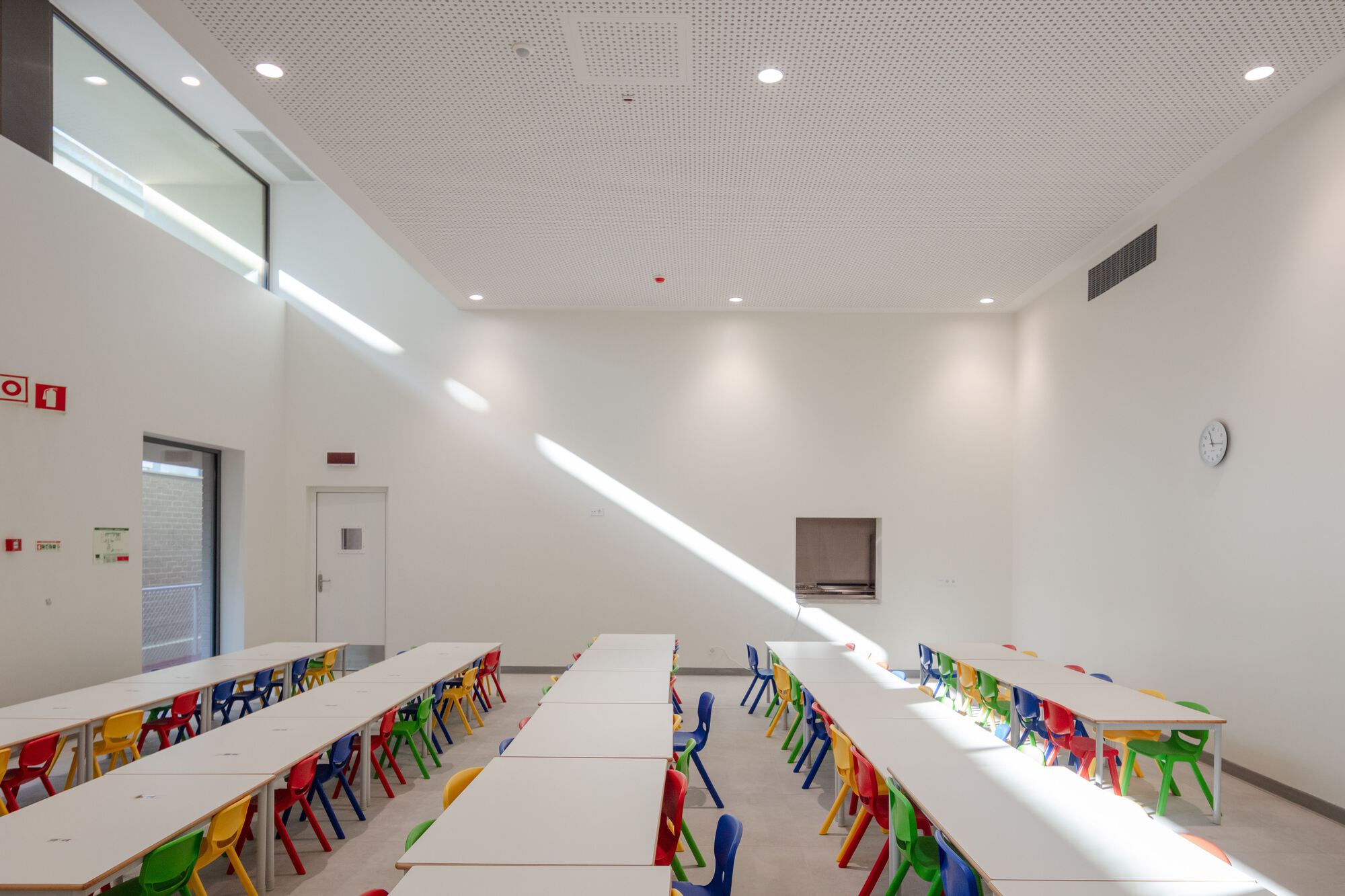 Dining hall with rows of white tables and bright plastic chairs illuminated by clerestory daylight