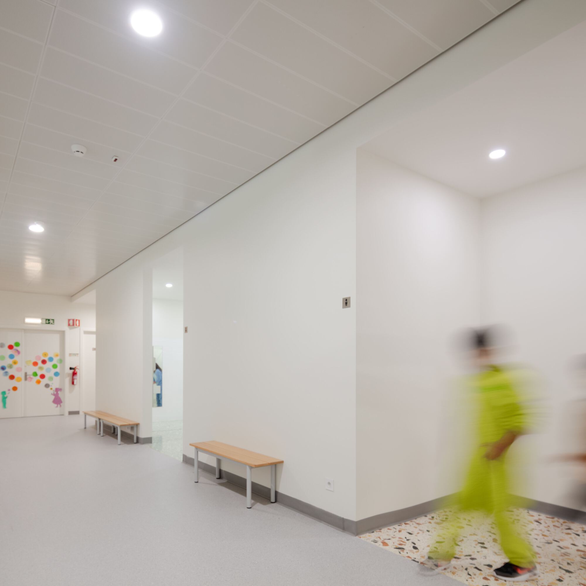 White hallway with terrazzo flooring and timber benches as a person in yellow walks past