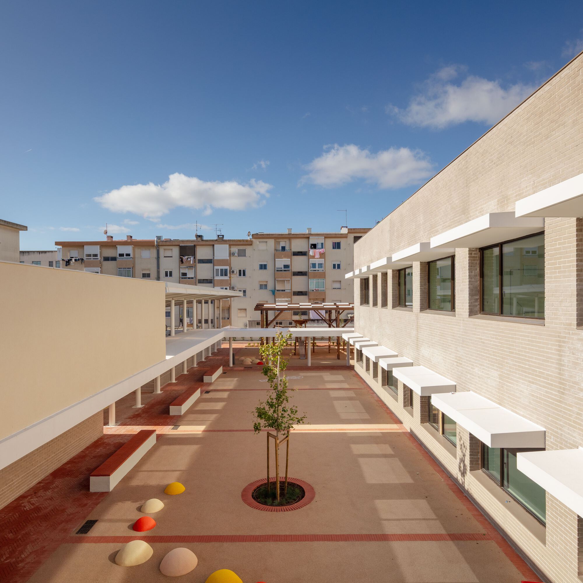 Central courtyard with a circular tree planter and scattered colored play spheres under partial clouds