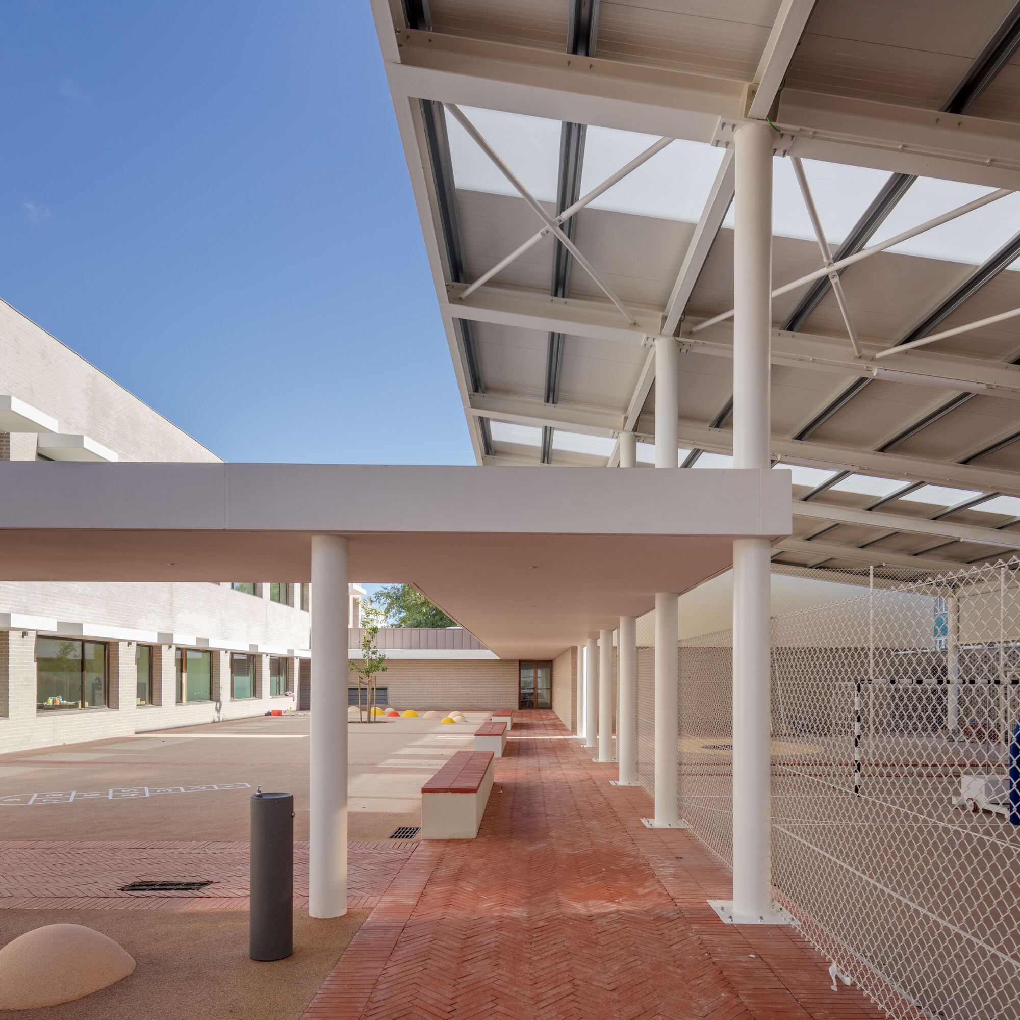 Covered walkway with white cylindrical columns supporting a cross-braced canopy over herringbone terracotta paving