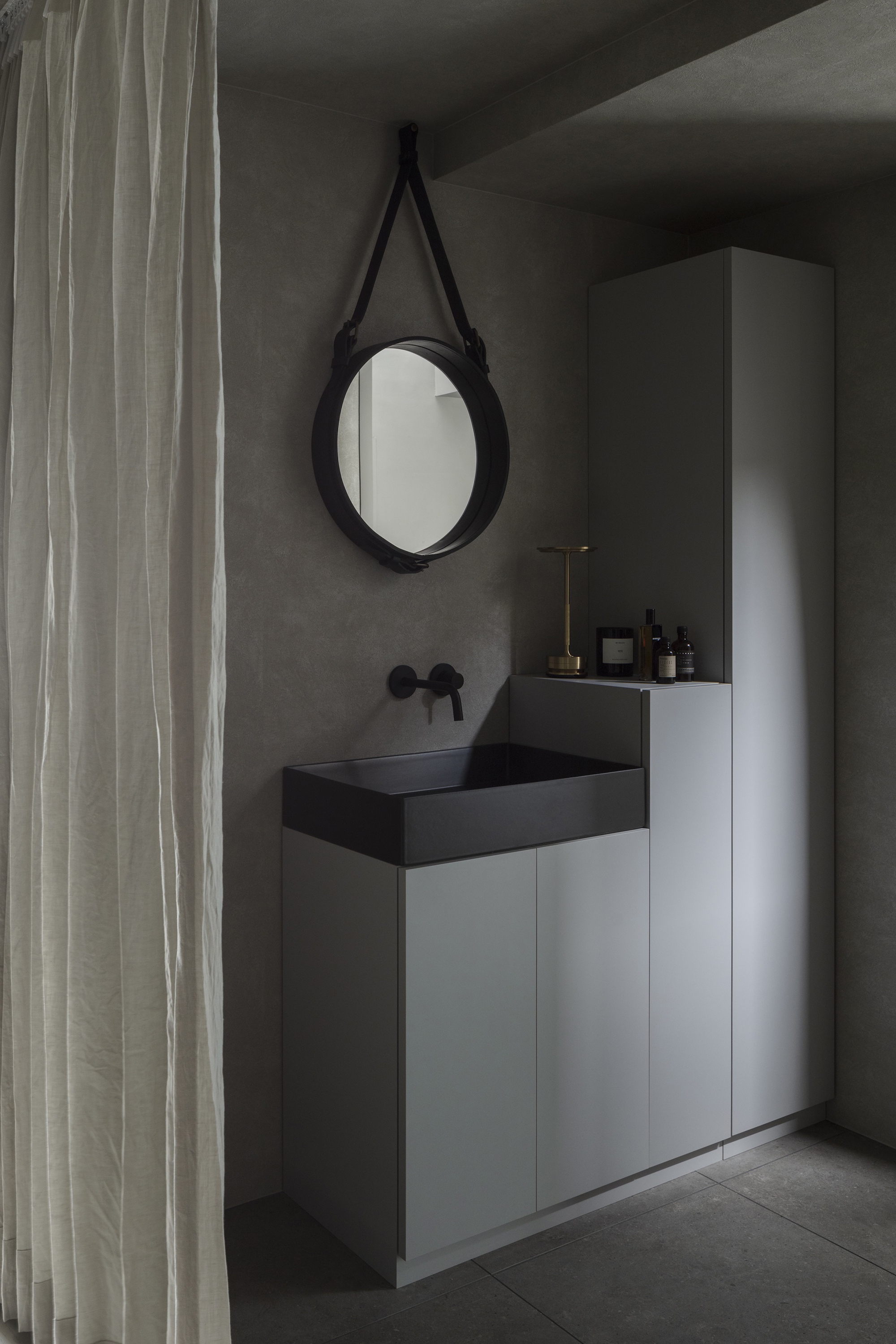 Bathroom vanity with black basin and round leather-strapped mirror beside linen curtain