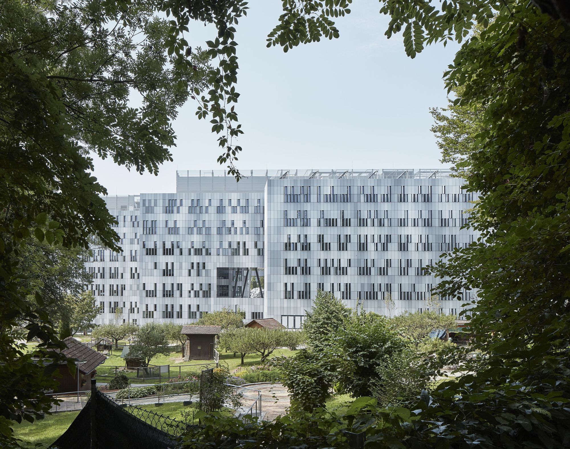White facade with vertical window pattern framed by overhanging tree branches and a garden plaza below
