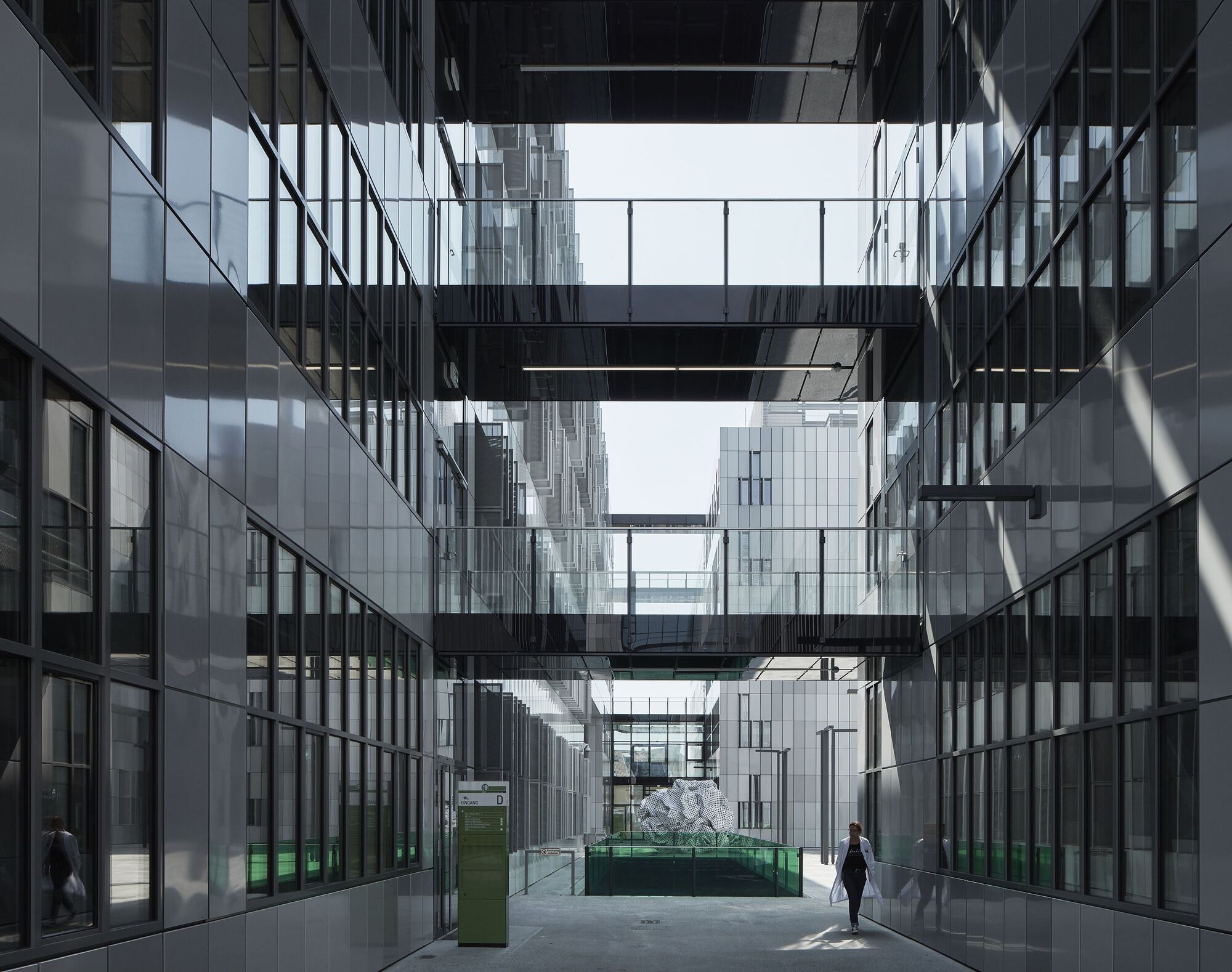 Glazed courtyard with black steel bridges connecting towers as a lone figure walks through sunlit space