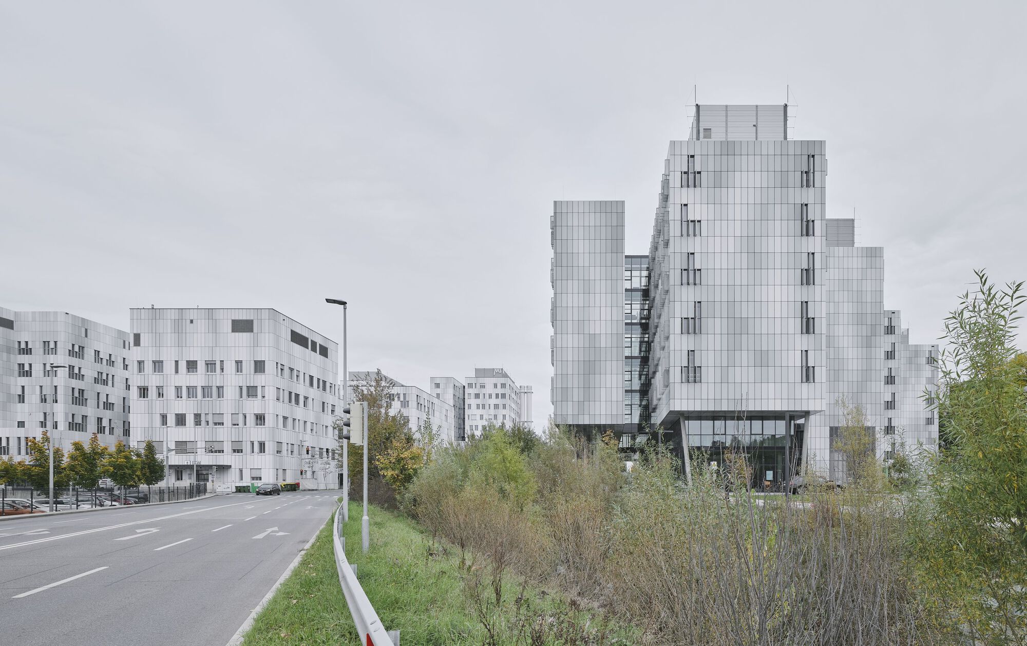 Stepped glass tower beside a roadway with wildflower meadow and residential blocks under overcast sky