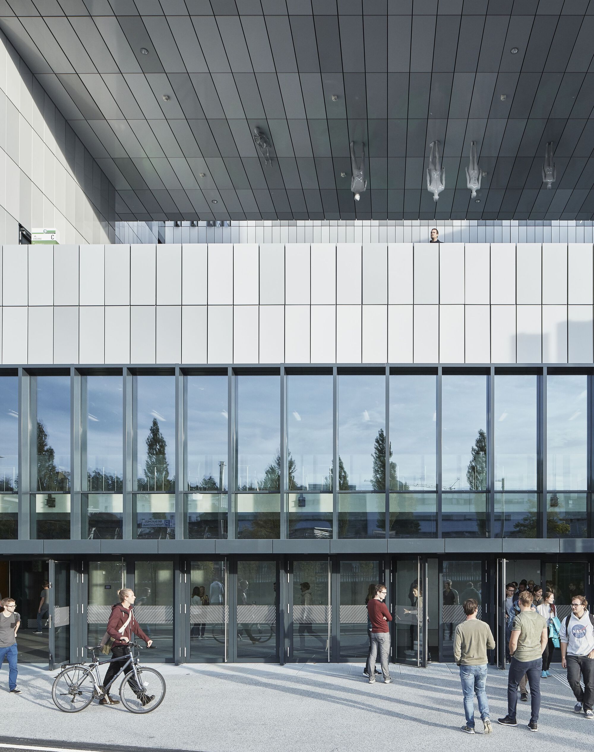 Glazed entrance facade with vertical white panels above and pedestrians gathering on the plaza