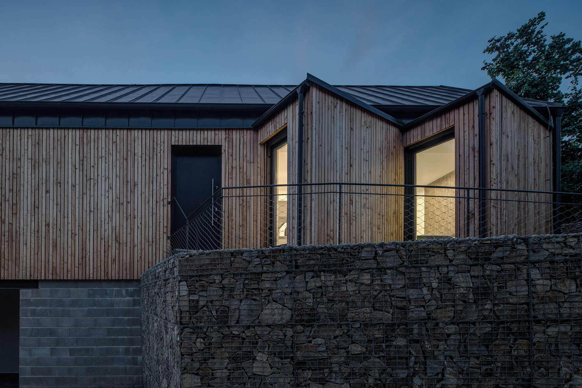 Vertical timber facade with illuminated windows at dusk above gabion retaining wall