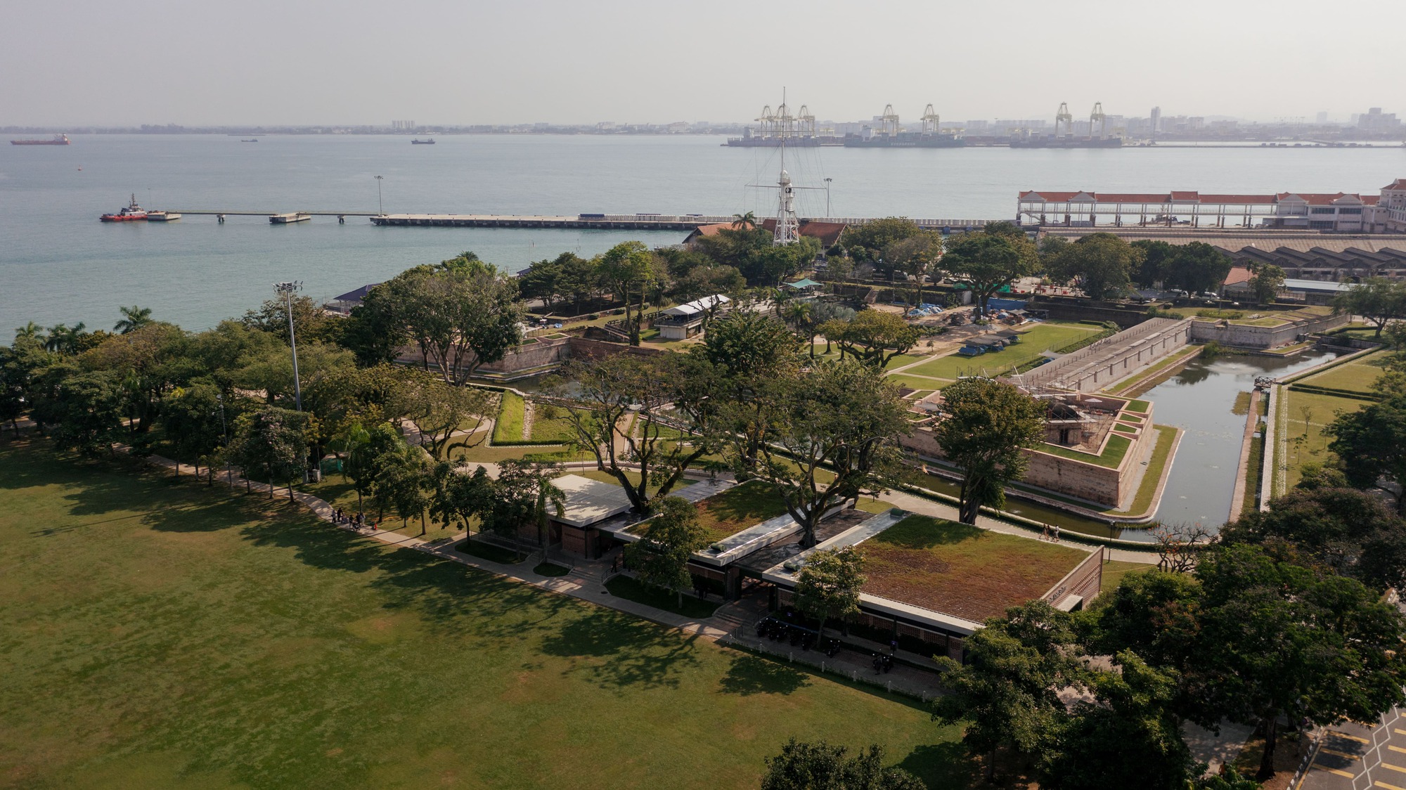 Aerial view of the pavilion complex with waterfront, harbor cranes, and park lawns in late afternoon light