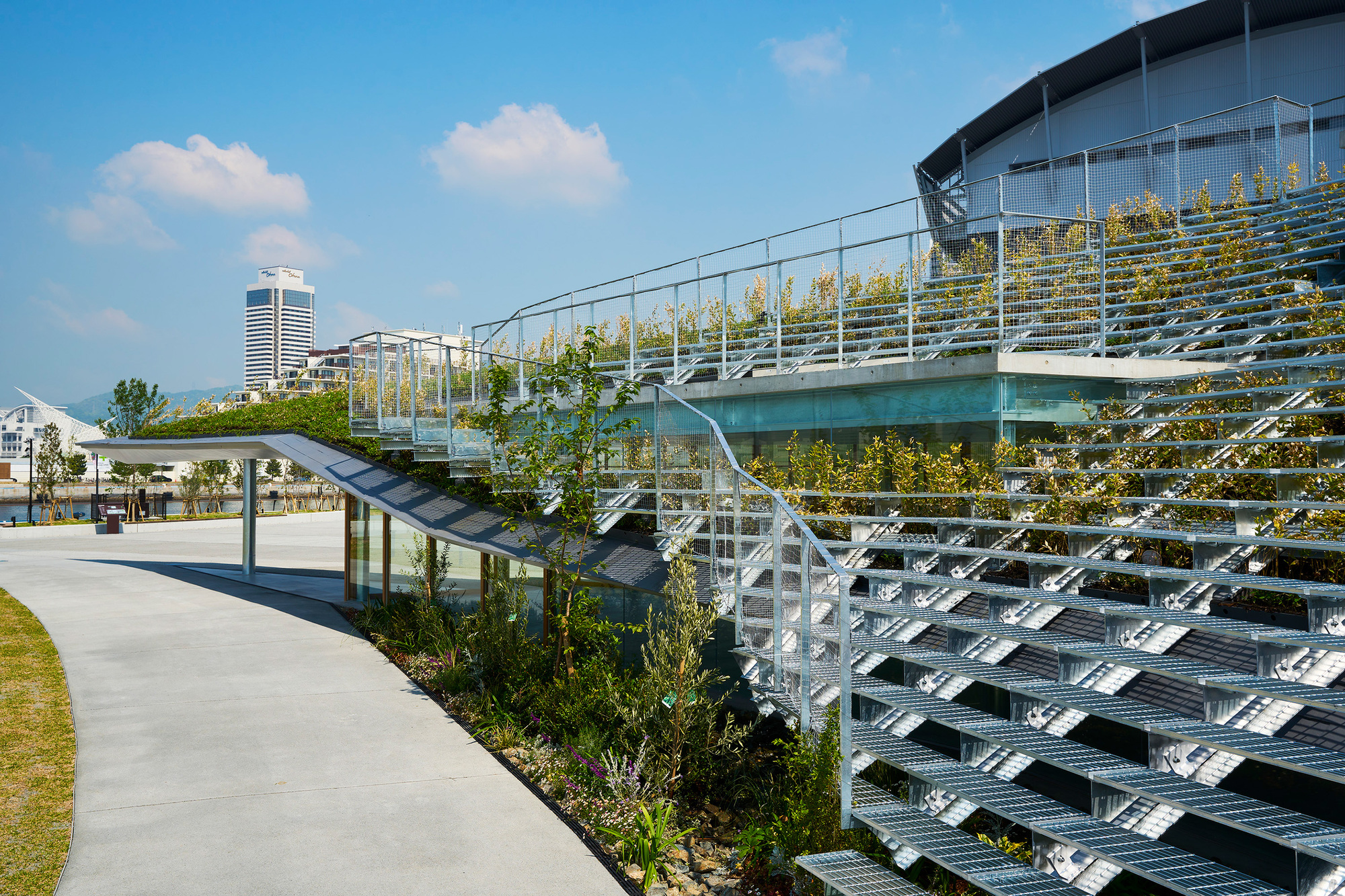 Metal grid terraces with planted rows alongside concrete path and pavilion under partly cloudy sky