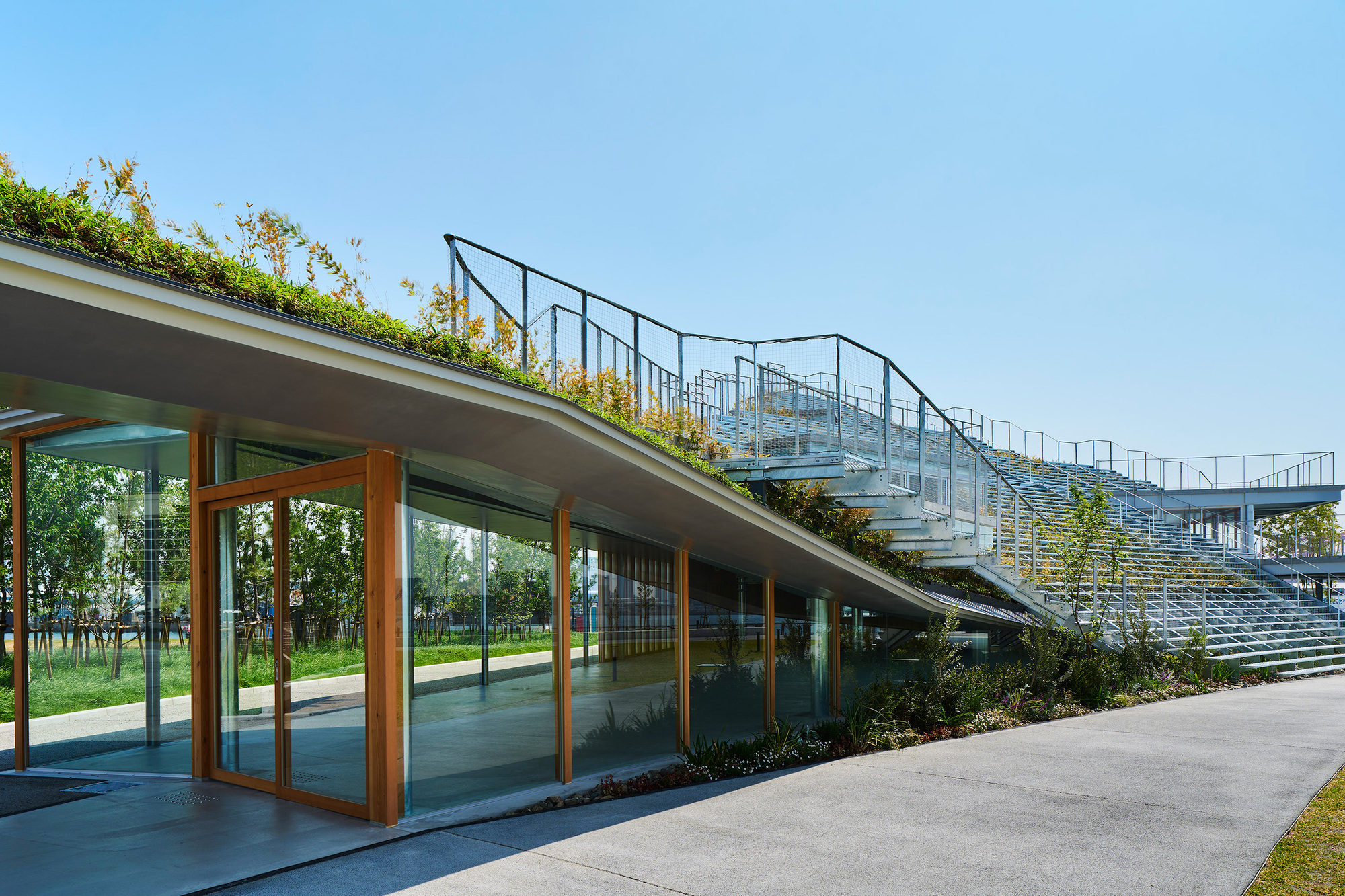Glass-fronted pavilion beneath planted roof and grandstand seating with metal railings under clear sky