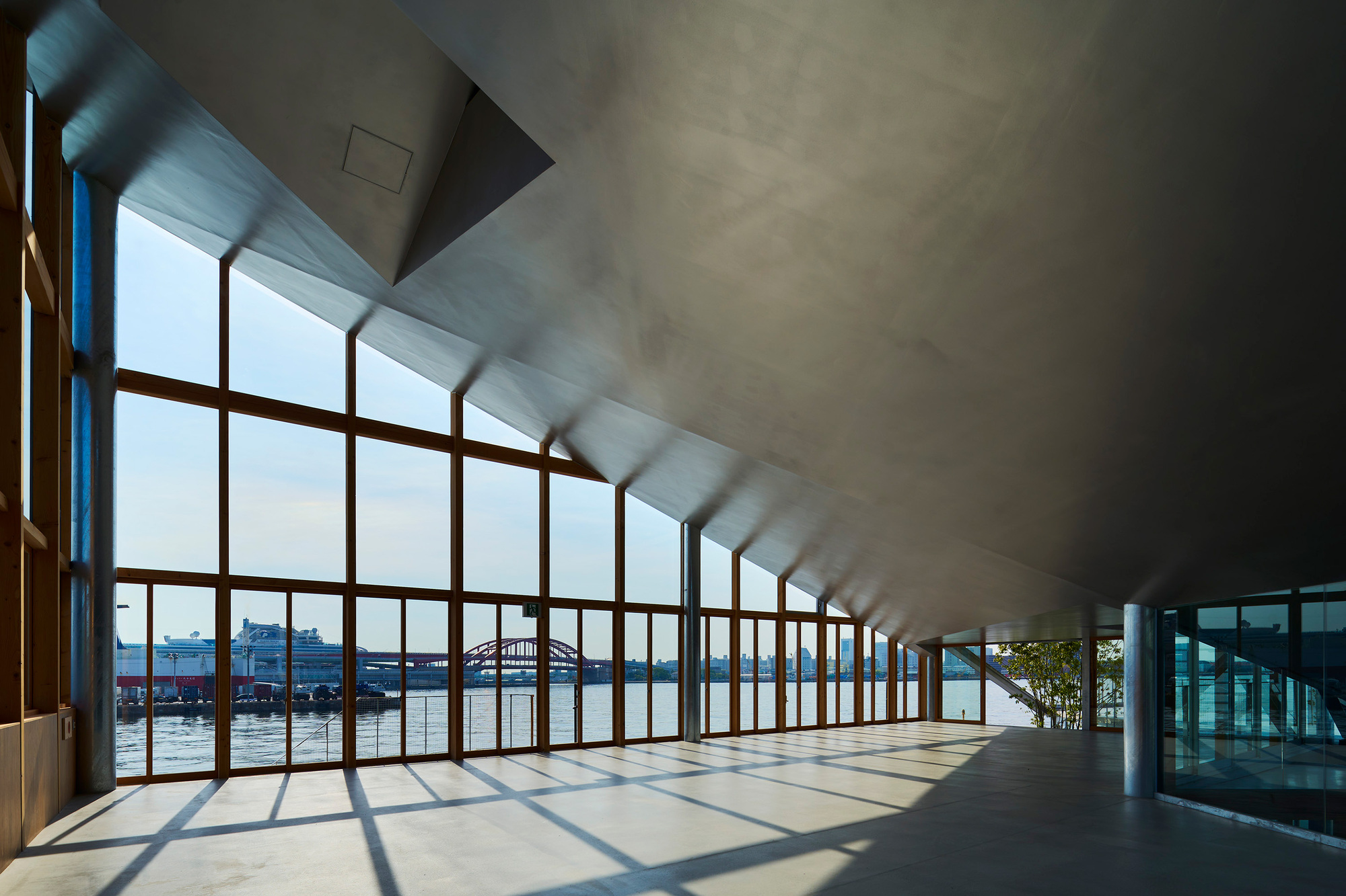 Sunlit corridor with timber curtain wall overlooking water and angled concrete ceiling plane above