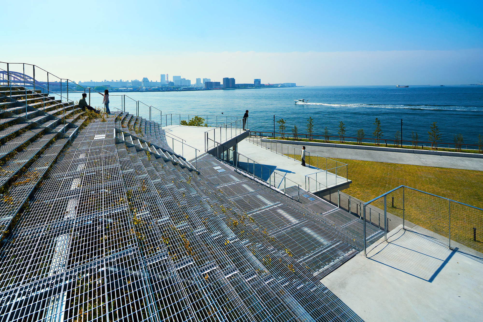 Tiered metal grate seating steps leading down to a waterfront lawn with visitors walking