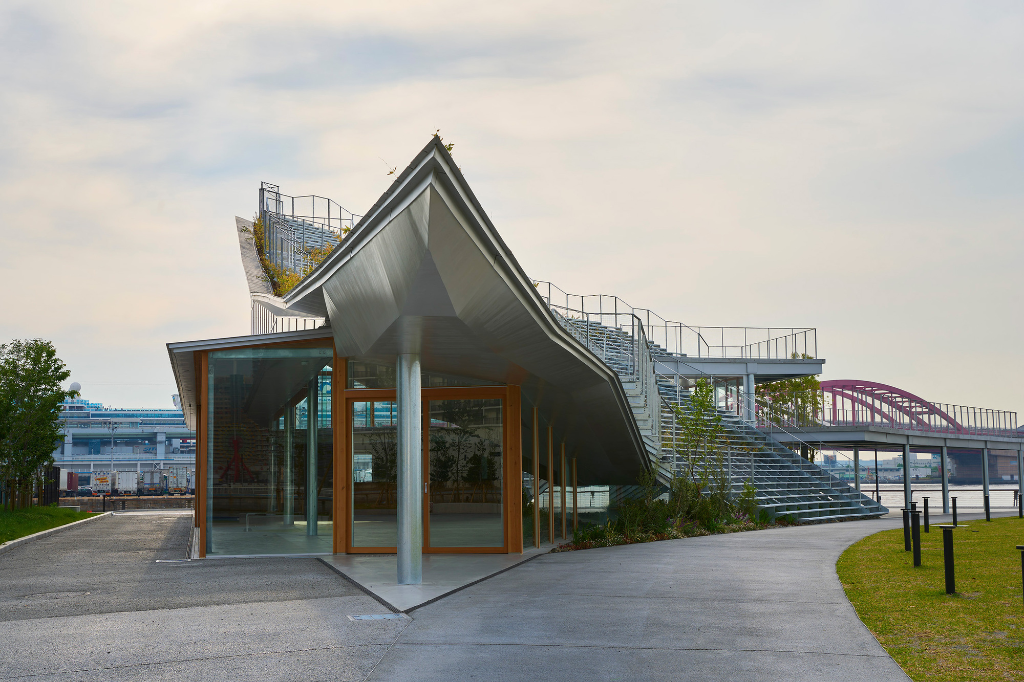 Glass pavilion with angular metal roof and external staircase beside planted lawn at dusk