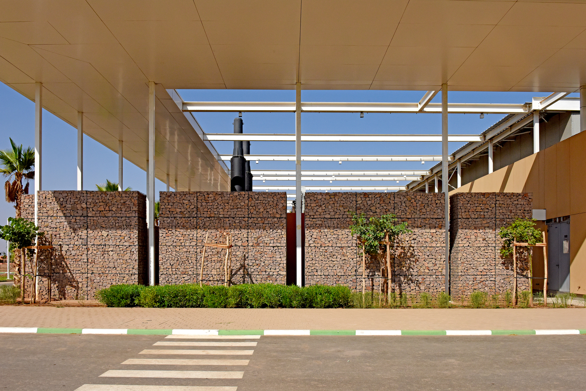 Gabion wall entry with white structural frame overhead and pedestrian crosswalk in front