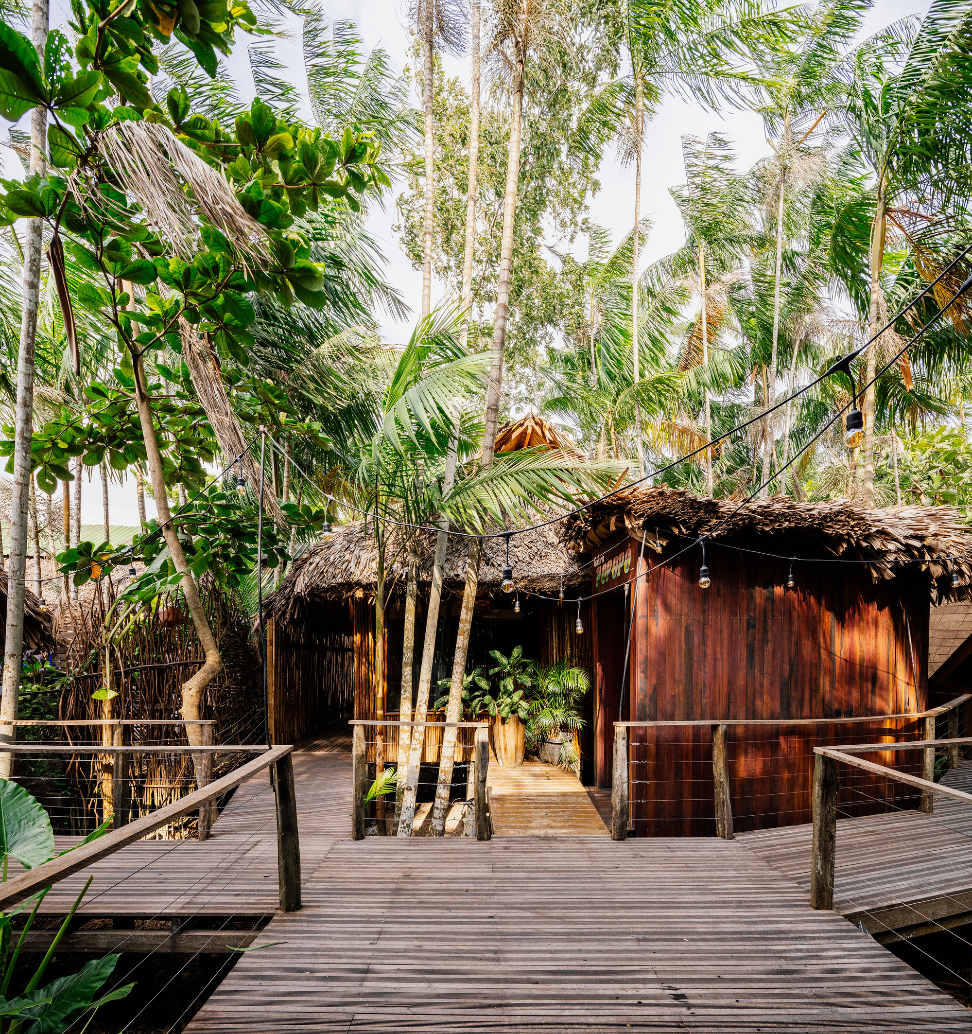 Timber boardwalk approaching thatched-roof pavilion with vertical wood cladding amid coconut palms