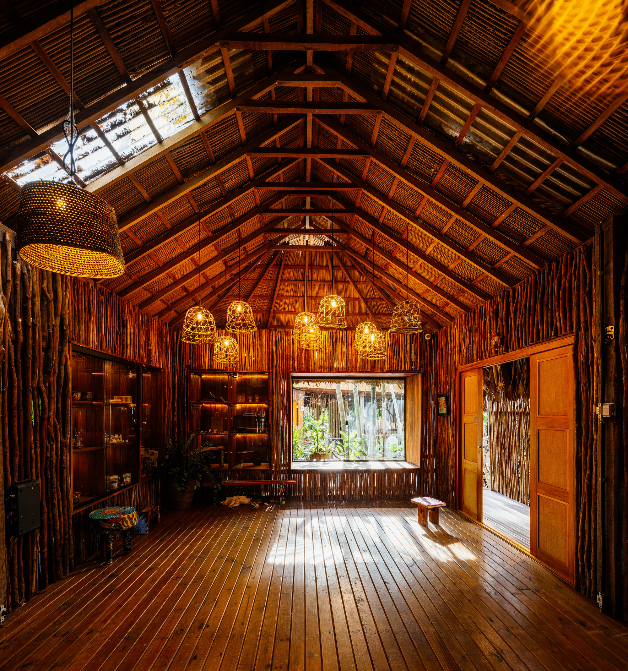 Interior hall with exposed timber trusses, woven pendant lights, and bamboo walls framing garden views