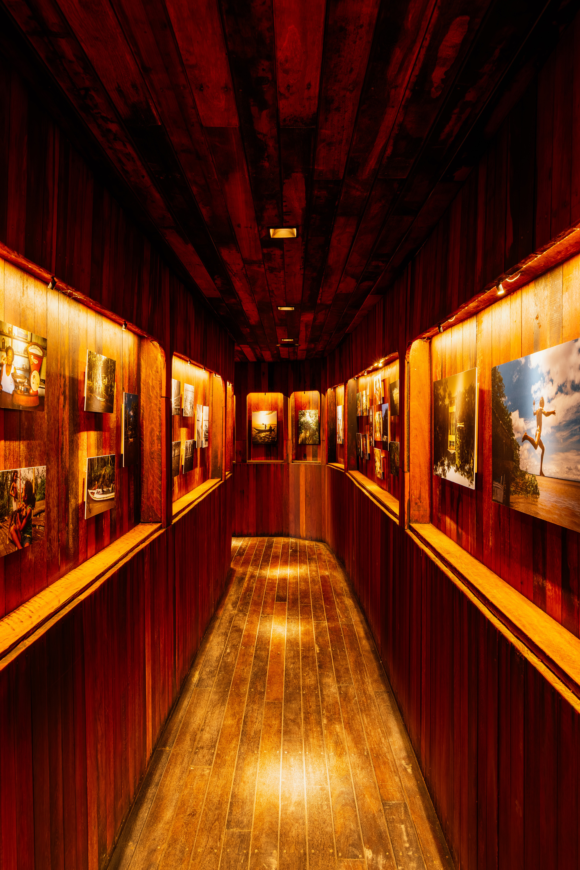 Gallery corridor with vertical timber walls displaying backlit photograph niches under exposed wood beams