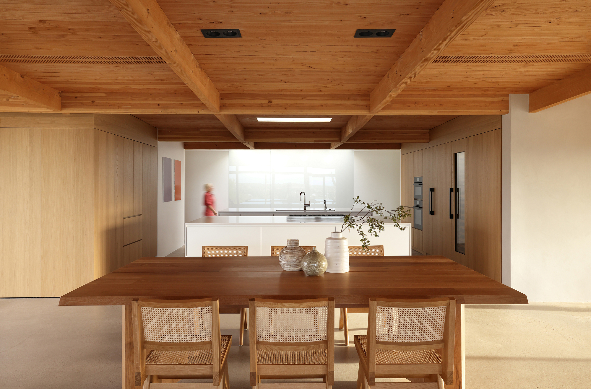 Dining area with timber table and caned chairs beneath heavy timber beams with person moving in kitchen beyond