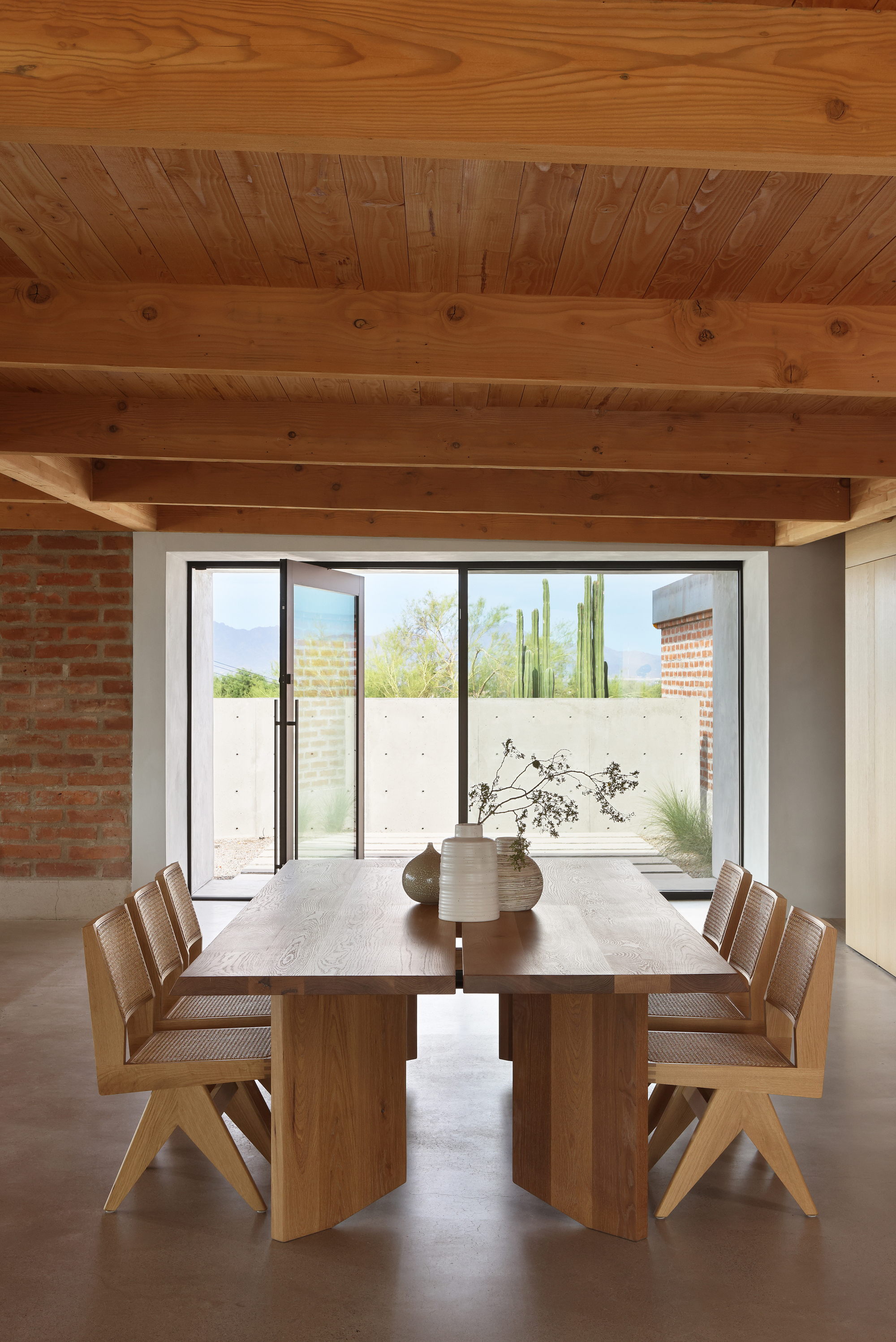 Dining table under exposed timber ceiling framed by glass doors opening to a desert courtyard