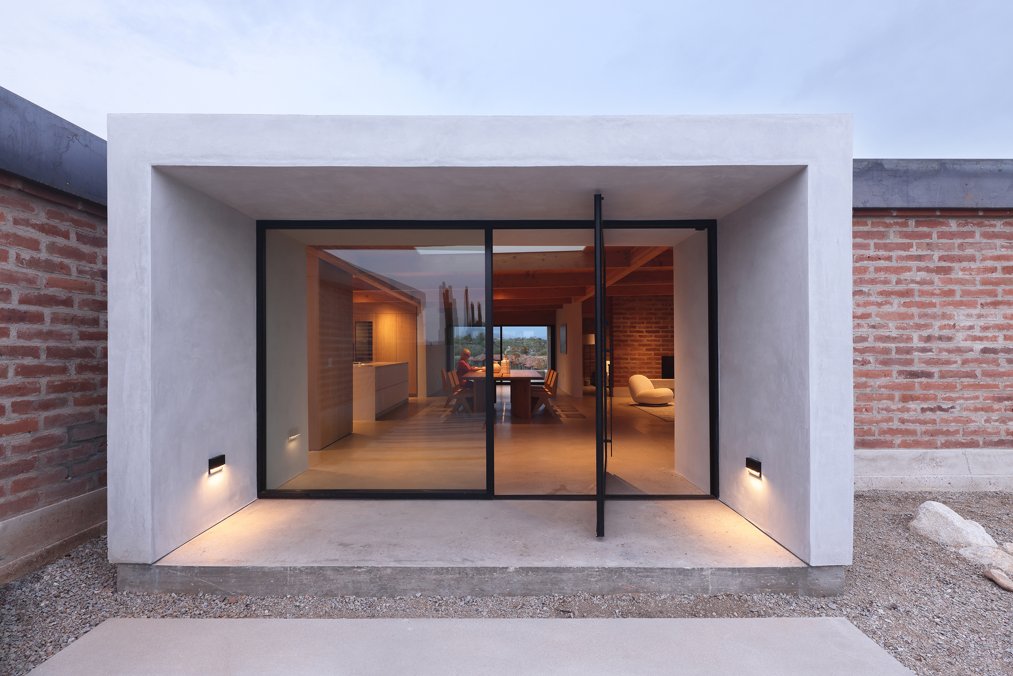 Entry pavilion with black steel pivot door, white stucco frame, and flanking brick walls at dusk