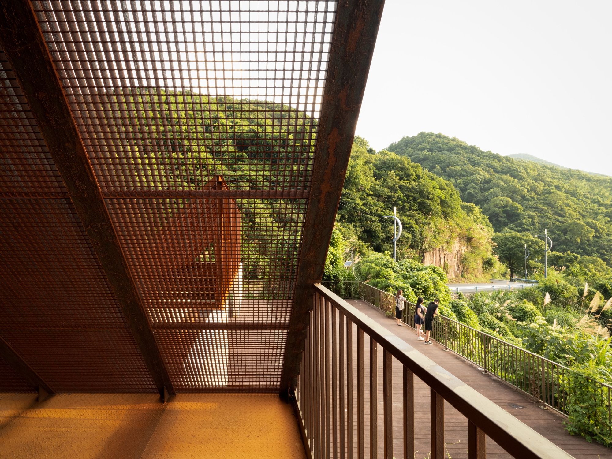 Interior balcony view through a corrugated metal mesh ceiling toward the forested hillside