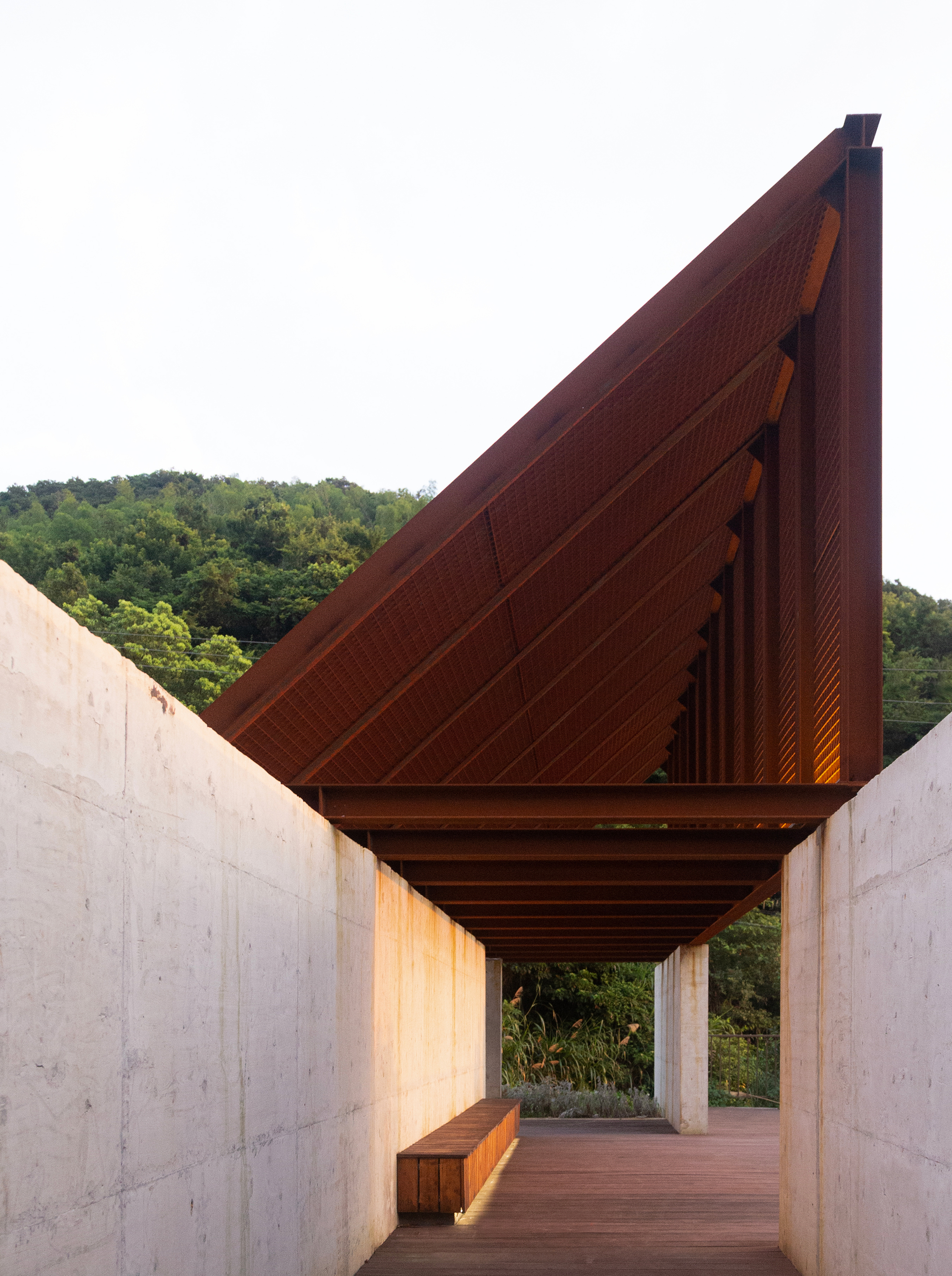 Covered walkway with exposed timber ceiling between concrete walls leading toward the hillside